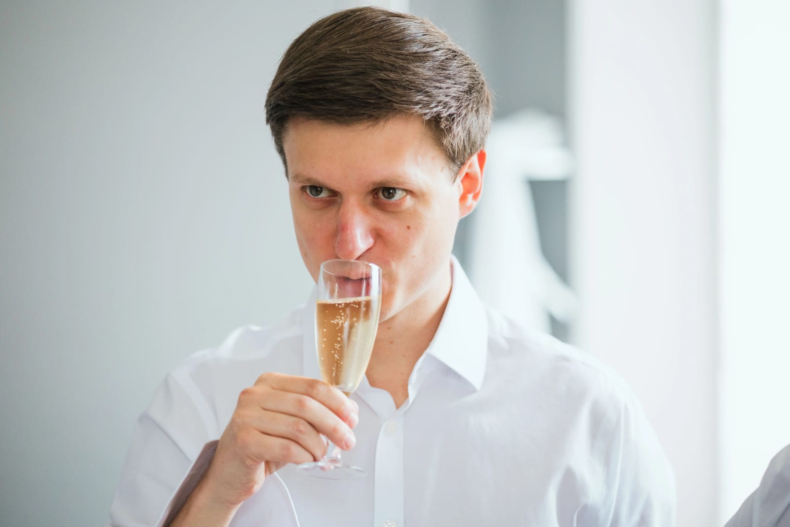 Un homme en chemise blanche boit une coupe de champagne en regardant l'objectif pendant les préparatifs, mariage au Domaine d'Orgival, par Jean-Baptiste Argyre (Jaysphotography)