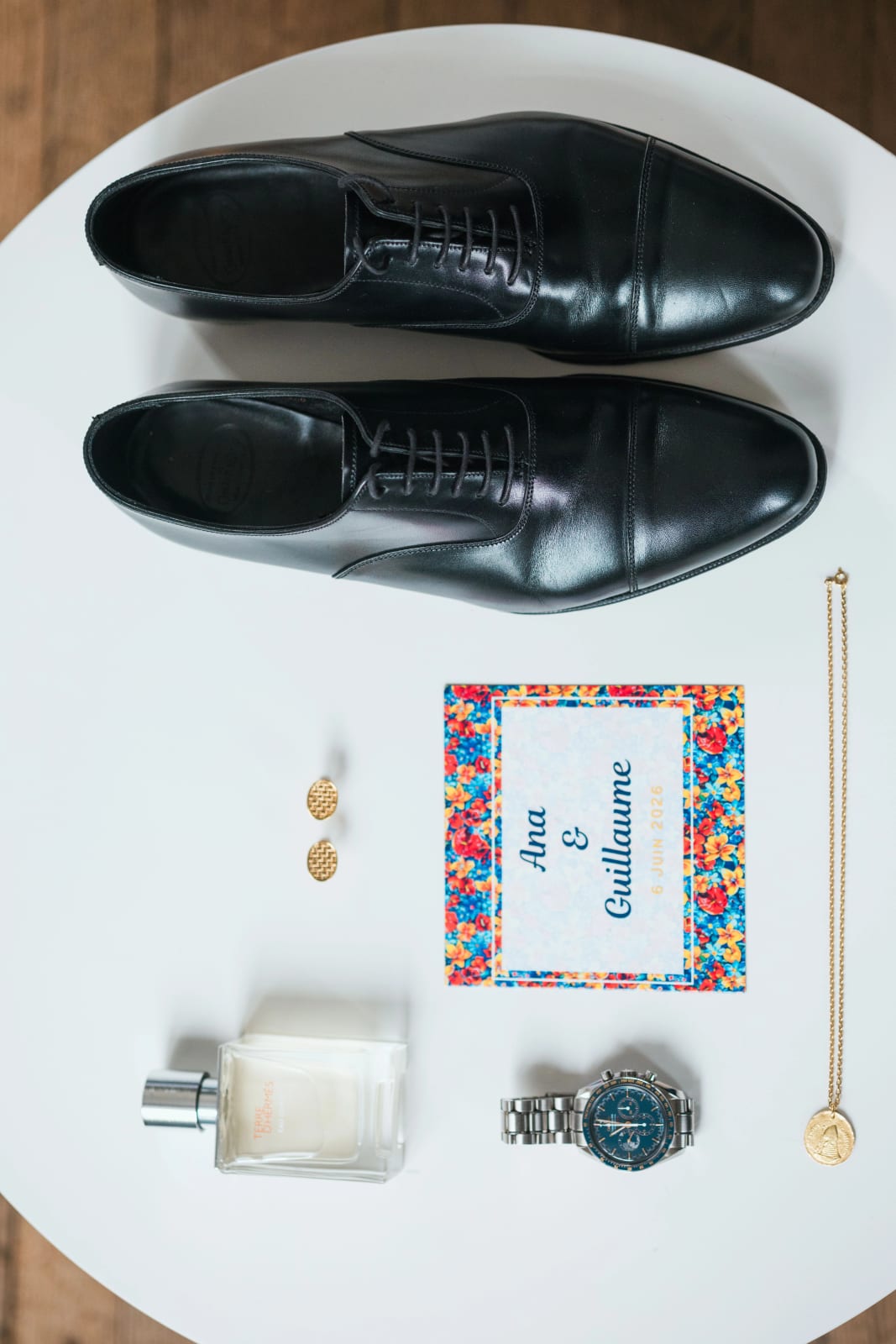 Mise à plat des accessoires du marié : souliers noirs, faire-part fleuri aux prénoms d'Ana et Guillaume, boutons de manchette dorés, parfum, montre, pendentif, mariage au Domaine d'Orgival, par Jean-Baptiste Argyre (Jaysphotography)