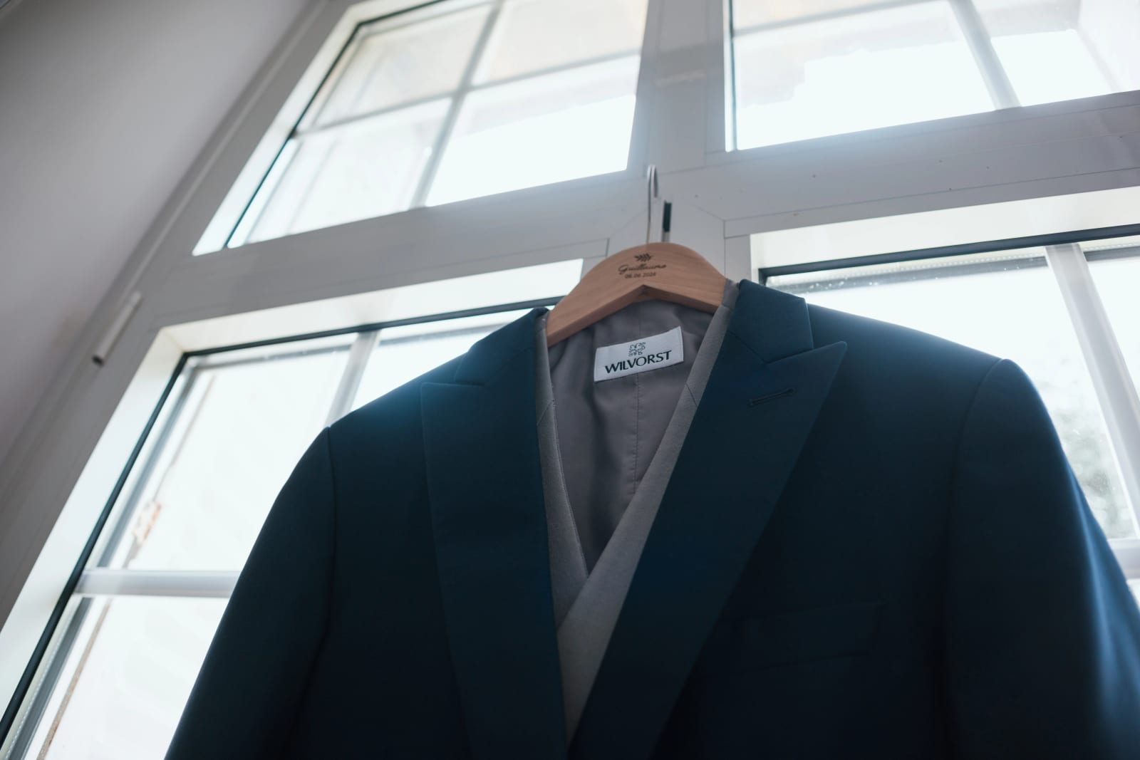 Le costume du marié, veste bleu canard à revers gris de la maison Wilvorst, suspendu à un cintre devant une fenêtre, mariage au Domaine d'Orgival, par Jean-Baptiste Argyre (Jaysphotography)