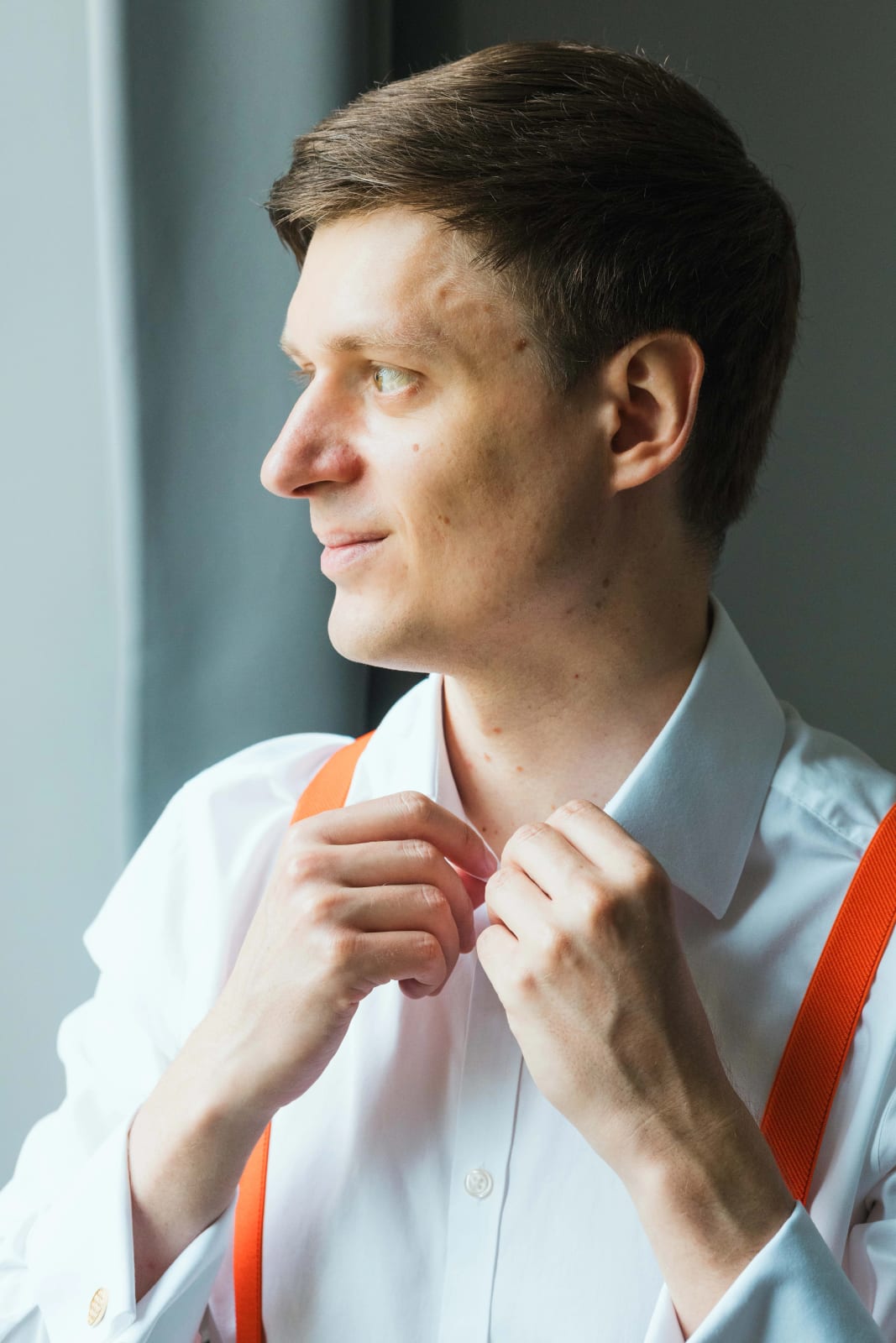 Guillaume, bretelles orange sur chemise blanche, ajuste son col en regardant par la fenêtre pendant son habillage, mariage au Domaine d'Orgival, par Jean-Baptiste Argyre (Jaysphotography)