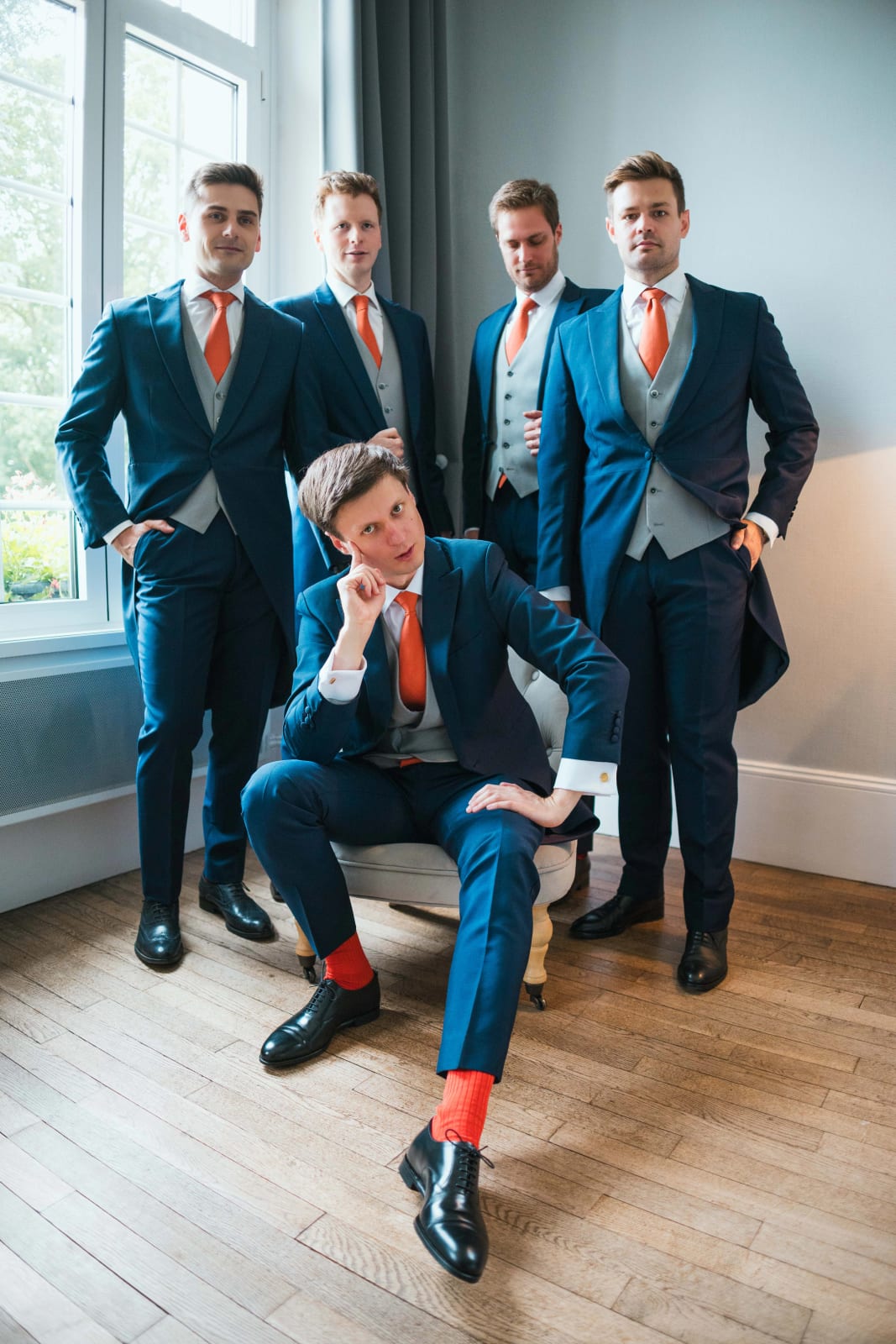 Guillaume assis au centre, chaussettes orange aux pieds, entouré de ses quatre garçons d'honneur en costume trois-pièces bleu et cravate orange, pose éditoriale dans un salon du château, mariage au Domaine d'Orgival, par Jean-Baptiste Argyre (Jaysphotography)
