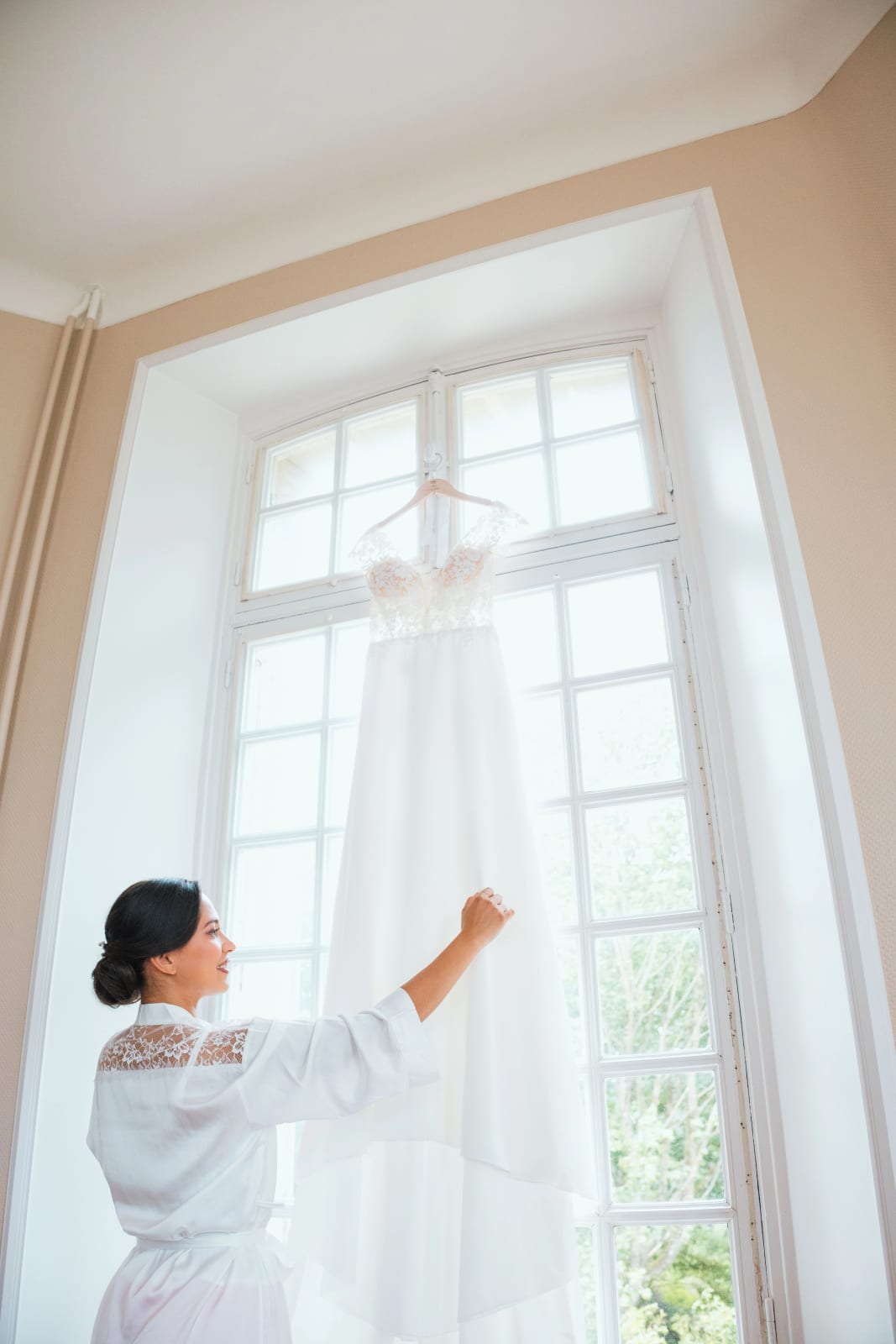 Ana de dos en peignoir clair tend la main vers sa robe de mariée suspendue à une haute fenêtre du château, mariage au Domaine d'Orgival, par Jean-Baptiste Argyre (Jaysphotography)