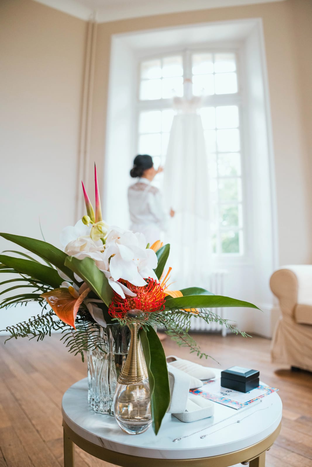 Bouquet tropical d'orchidées blanches, d'anthuriums orange et de palmes posé sur une table, Ana floue à l'arrière près de sa robe, mariage au Domaine d'Orgival, par Jean-Baptiste Argyre (Jaysphotography)