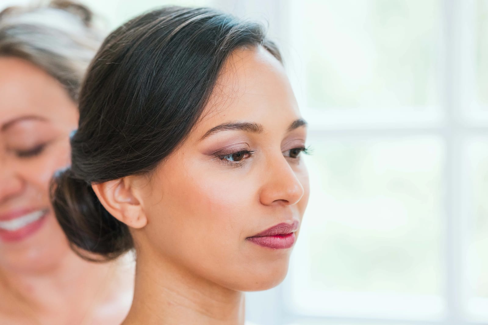 Portrait rapproché d'Ana maquillée, regard baissé, chignon bas, une proche souriante en arrière-plan, mariage au Domaine d'Orgival, par Jean-Baptiste Argyre (Jaysphotography)