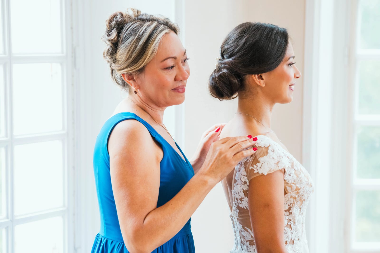 Une proche en robe bleu roi ferme le haut en dentelle de la robe de mariée sur l'épaule d'Ana, mariage au Domaine d'Orgival, par Jean-Baptiste Argyre (Jaysphotography)