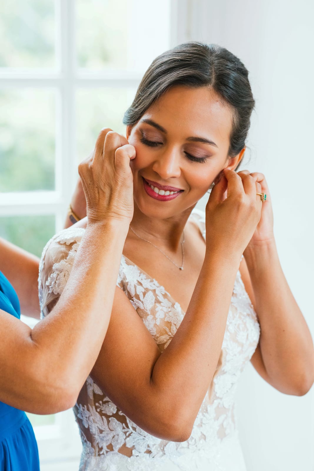 Ana, yeux fermés et sourire aux lèvres, met ses boucles d'oreilles dans sa robe en dentelle, aidée par une proche, mariage au Domaine d'Orgival, par Jean-Baptiste Argyre (Jaysphotography)