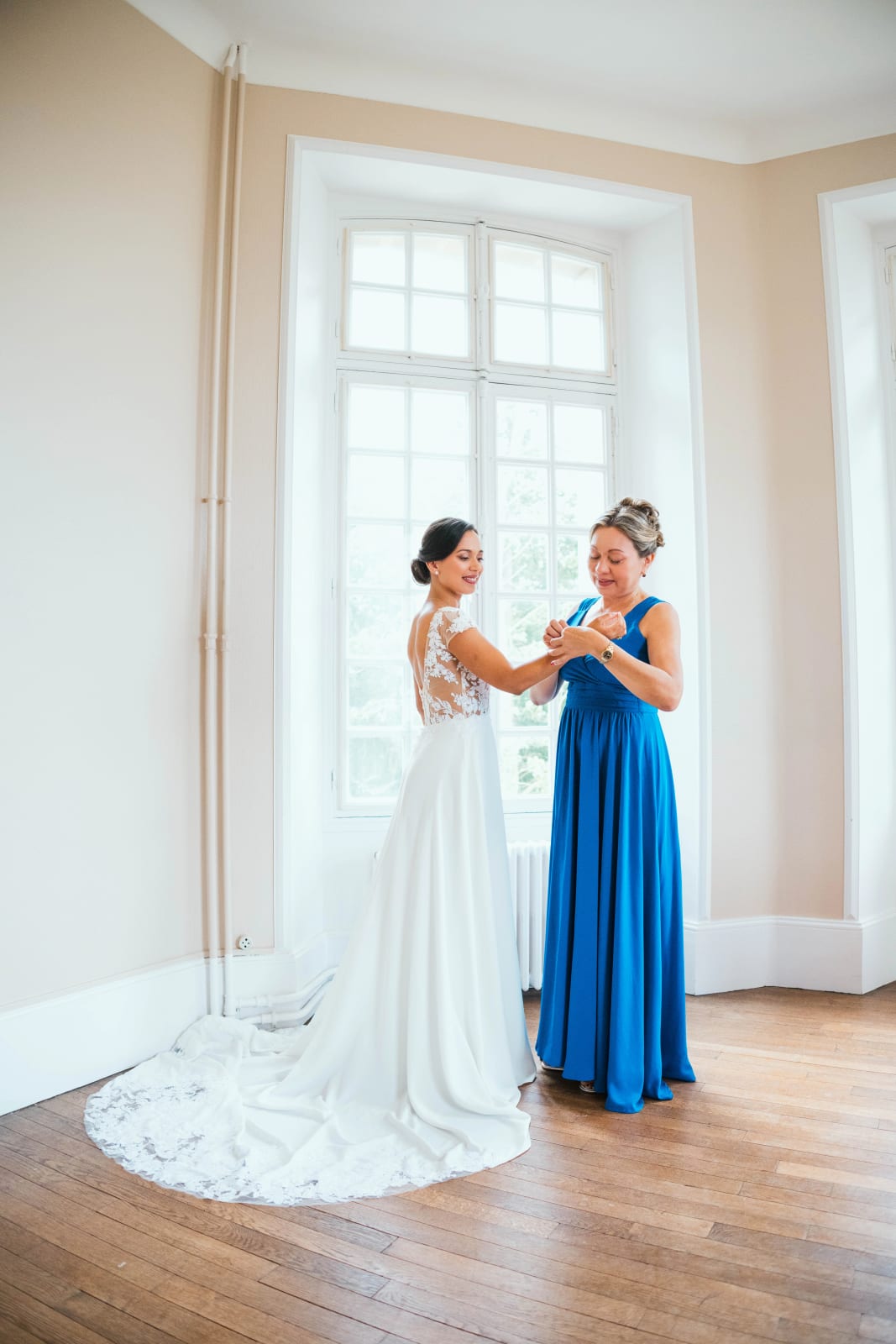Ana en robe de mariée à dos de dentelle et longue traîne, face à une haute fenêtre, une proche en robe bleu roi à ses côtés, dans un salon du château, mariage au Domaine d'Orgival, par Jean-Baptiste Argyre (Jaysphotography)