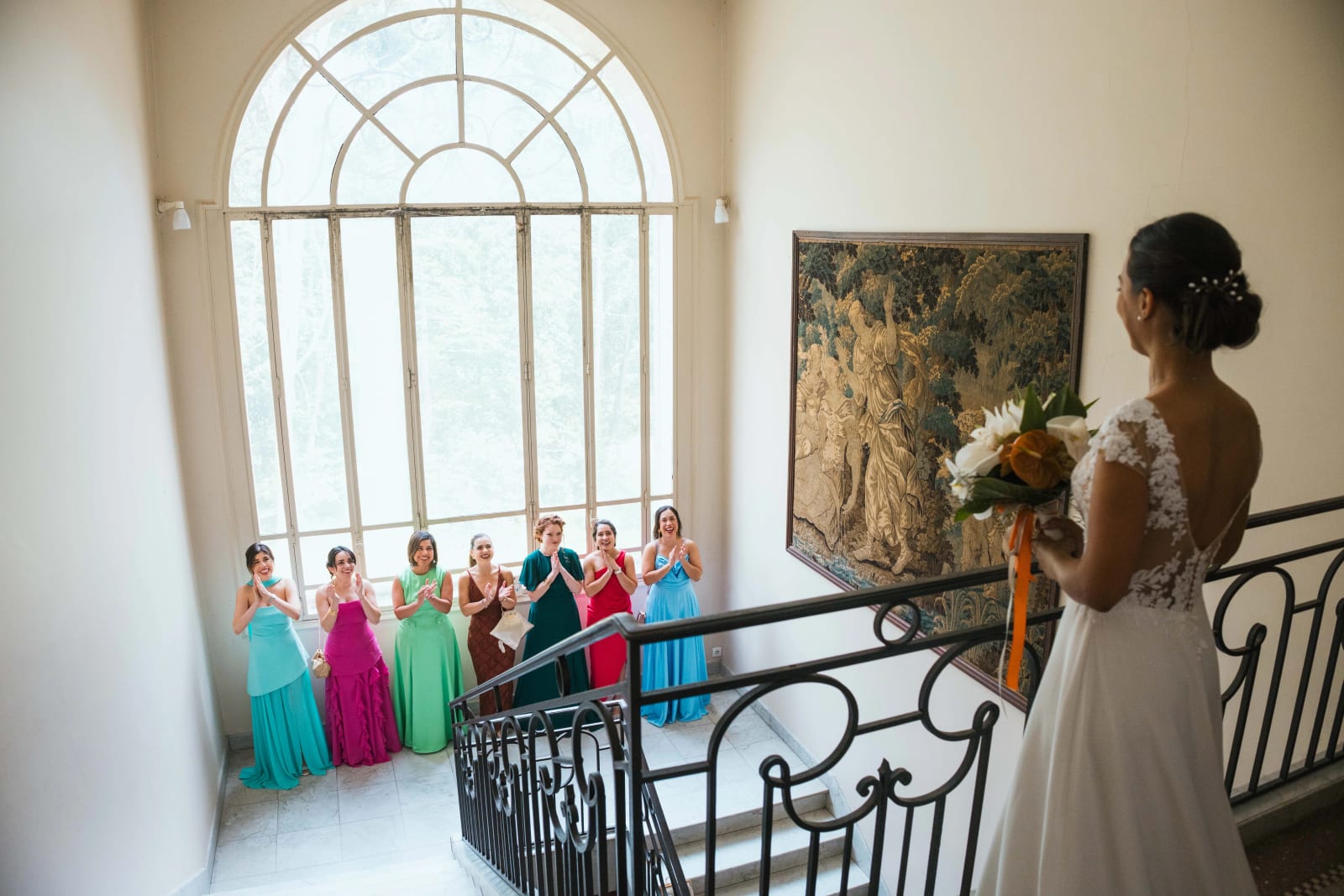 Ana de dos, bouquet à la main, se dévoile à ses demoiselles d'honneur en robes longues colorées réunies au pied du grand escalier du château, mariage au Domaine d'Orgival, par Jean-Baptiste Argyre (Jaysphotography)