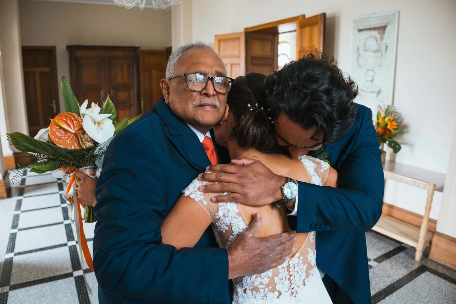 Ana enlacée par son papa très ému et par un proche, instant d'émotion dans un salon du château, bouquet tropical au premier plan, mariage au Domaine d'Orgival, par Jean-Baptiste Argyre (Jaysphotography)