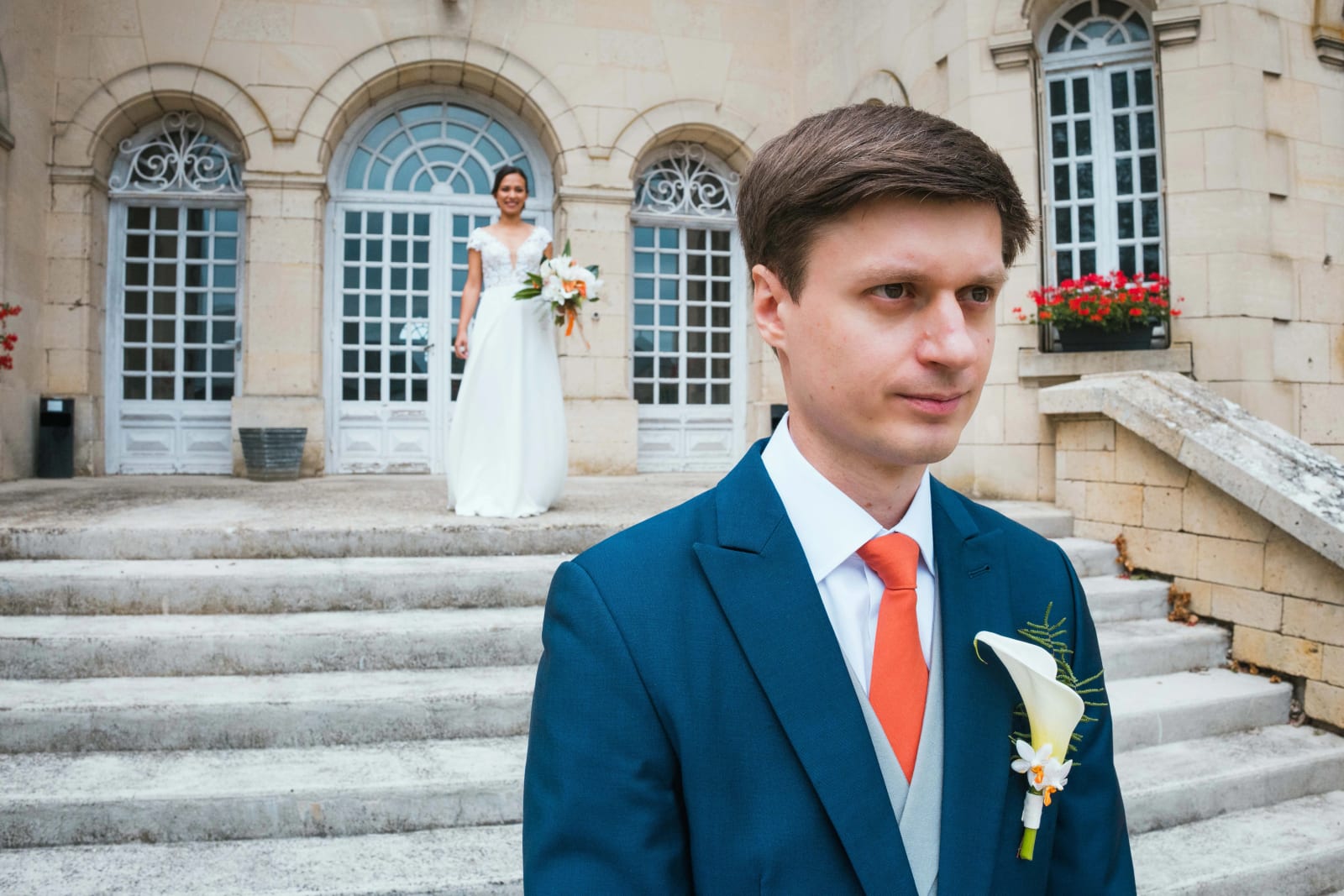 First look : Guillaume en costume bleu et cravate orange au premier plan, Ana floue derrière lui sur le perron du château aux portes cintrées, mariage au Domaine d'Orgival, par Jean-Baptiste Argyre (Jaysphotography)