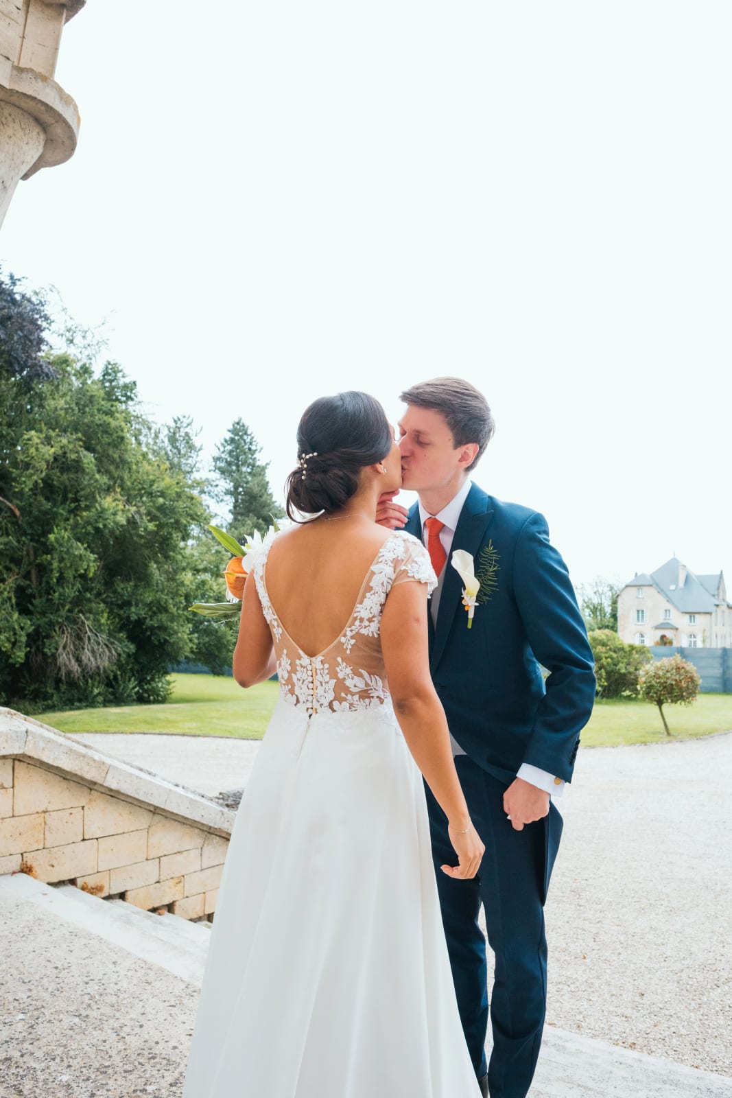 Ana, dos en dentelle face à l'objectif, embrasse Guillaume après le first look, un château en arrière-plan dans la verdure, mariage au Domaine d'Orgival, par Jean-Baptiste Argyre (Jaysphotography)