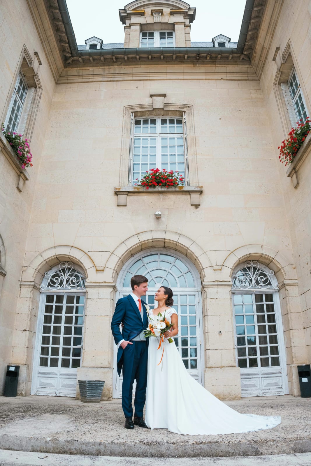 Ana et Guillaume enlacés au pied de la façade en pierre du château, hautes portes vitrées et jardinières de géraniums rouges, longue traîne déployée, mariage au Domaine d'Orgival, par Jean-Baptiste Argyre (Jaysphotography)