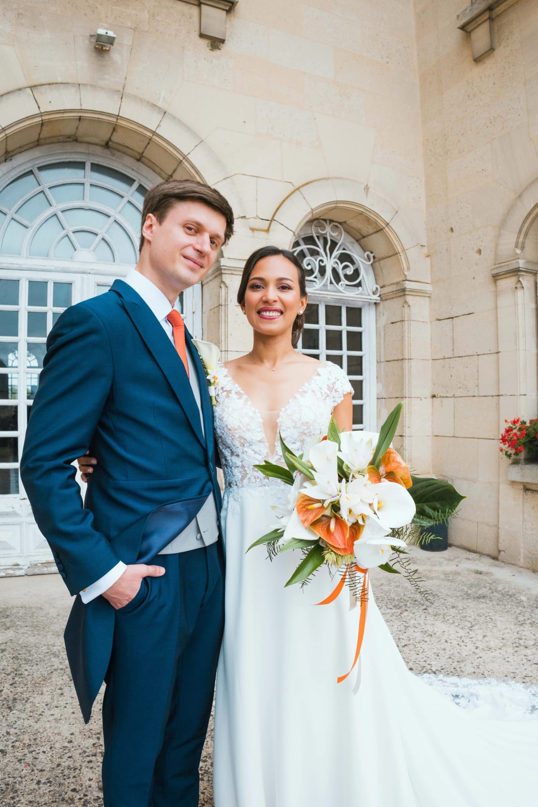 Portrait d'Ana et Guillaume souriants devant les arcs en pierre du château, Ana tenant son grand bouquet tropical blanc et orange, mariage au Domaine d'Orgival, par Jean-Baptiste Argyre (Jaysphotography)
