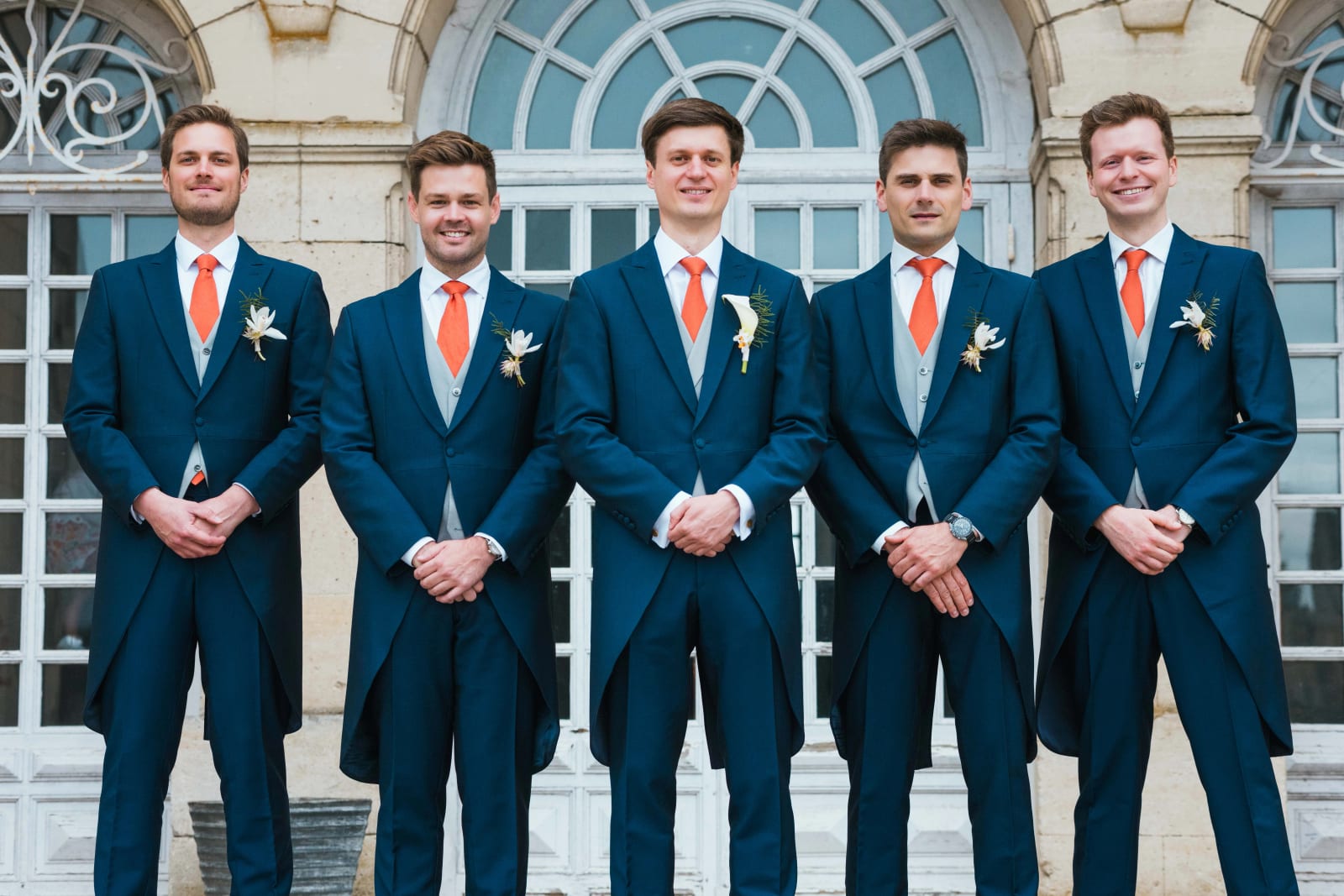 Les cinq hommes, Guillaume et ses garçons d'honneur, en costume bleu, gilet gris et cravate orange, alignés devant les fenêtres cintrées du château, mariage au Domaine d'Orgival, par Jean-Baptiste Argyre (Jaysphotography)