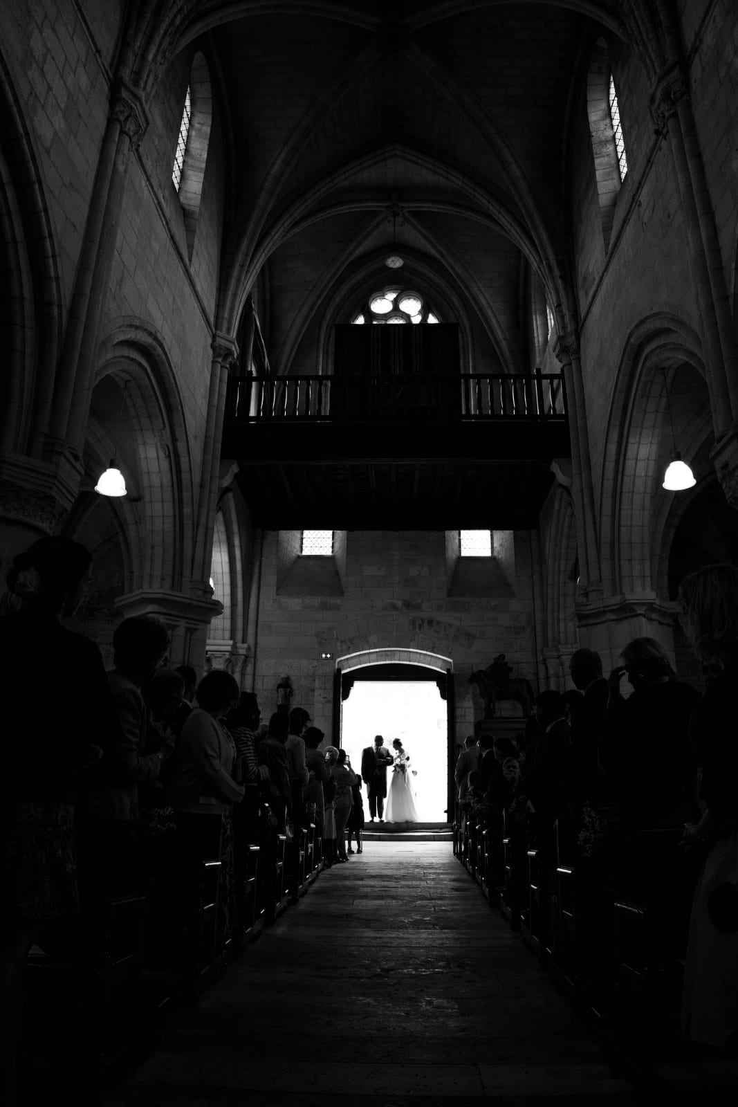 Vue en noir et blanc de la nef romane de l'église d'Ambleny, les mariés silhouettés à contre-jour dans le portail, invités dans les bancs, mariage au Domaine d'Orgival, par Jean-Baptiste Argyre (Jaysphotography)