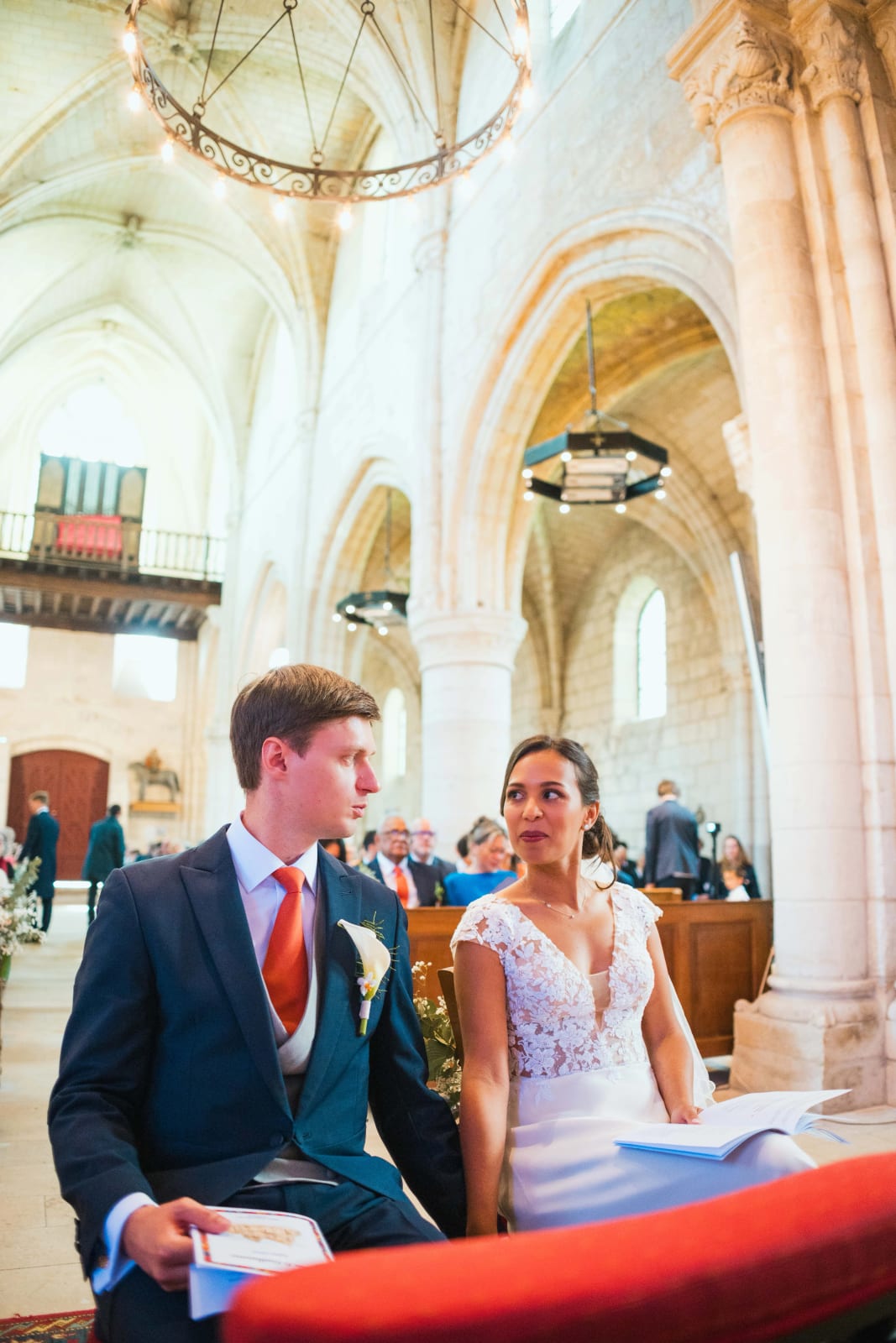 Ana et Guillaume assis côte à côte pendant la cérémonie religieuse, livrets bleus en main, sous les voûtes de pierre de l'église d'Ambleny, mariage au Domaine d'Orgival, par Jean-Baptiste Argyre (Jaysphotography)