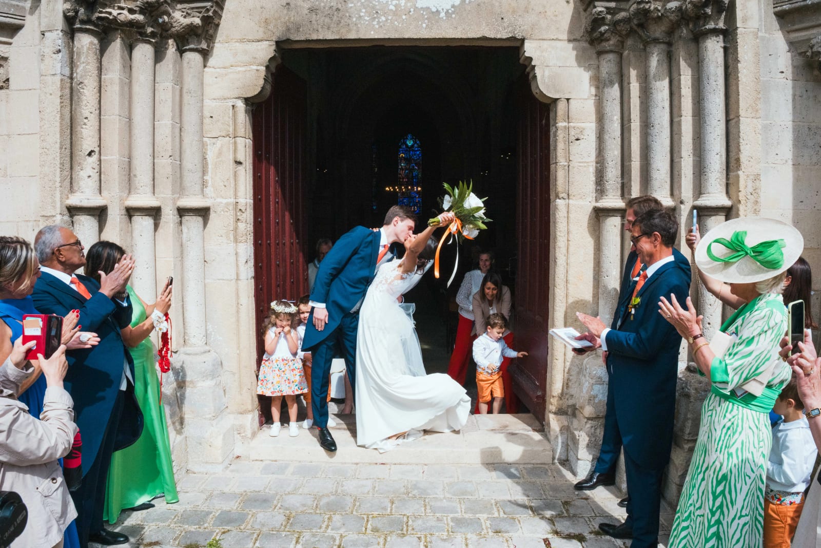 Sortie de l'église d'Ambleny : Ana et Guillaume s'embrassent sur le seuil sous une pluie de confettis, invités applaudissant, enfants d'honneur dans l'encadrement, mariage au Domaine d'Orgival, par Jean-Baptiste Argyre (Jaysphotography)