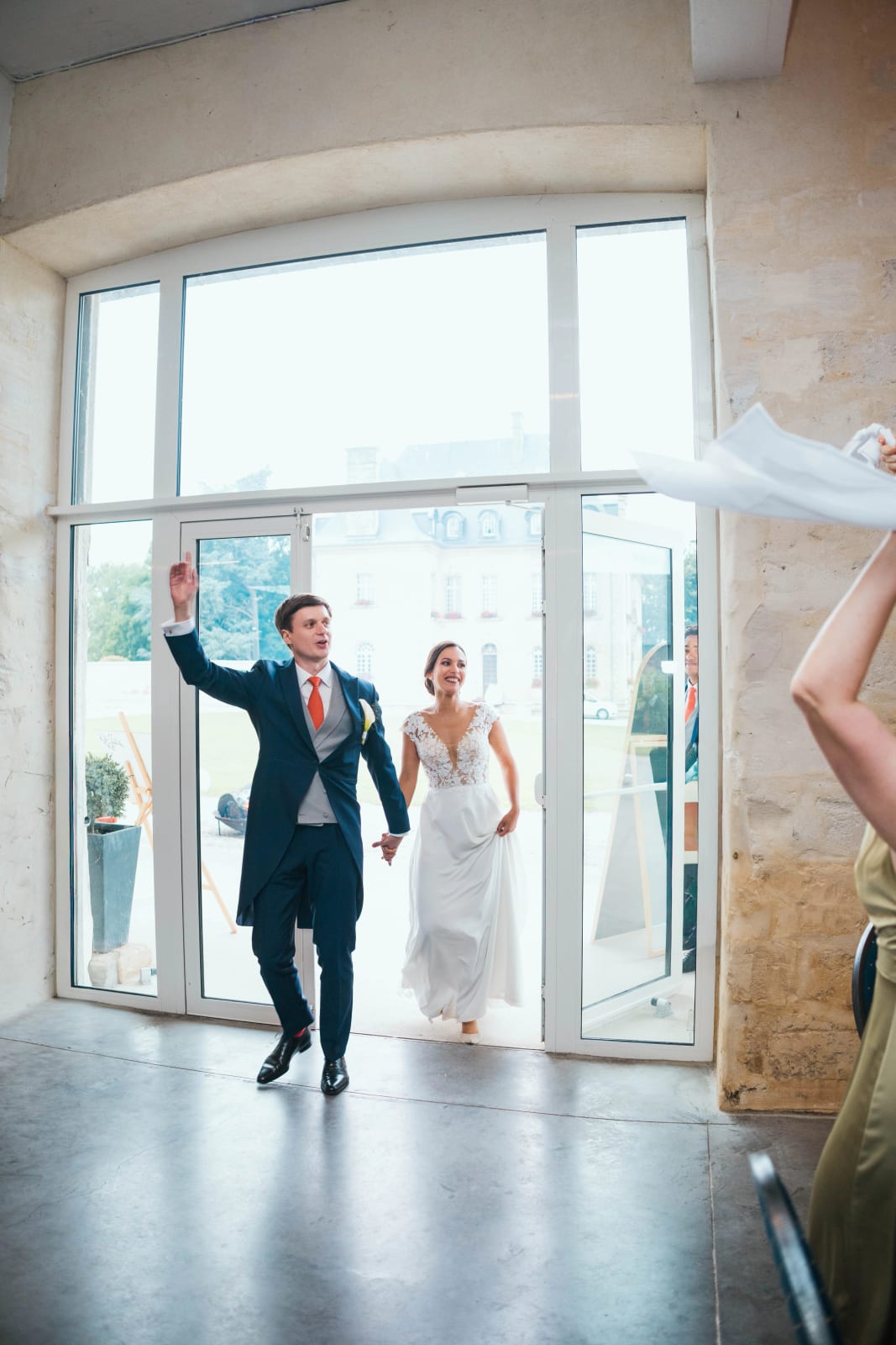 Entrée en salle des mariés : Guillaume mène Ana par la main à travers les portes vitrées, le château visible dehors, une invitée agitant une serviette, mariage au Domaine d'Orgival, par Jean-Baptiste Argyre (Jaysphotography)