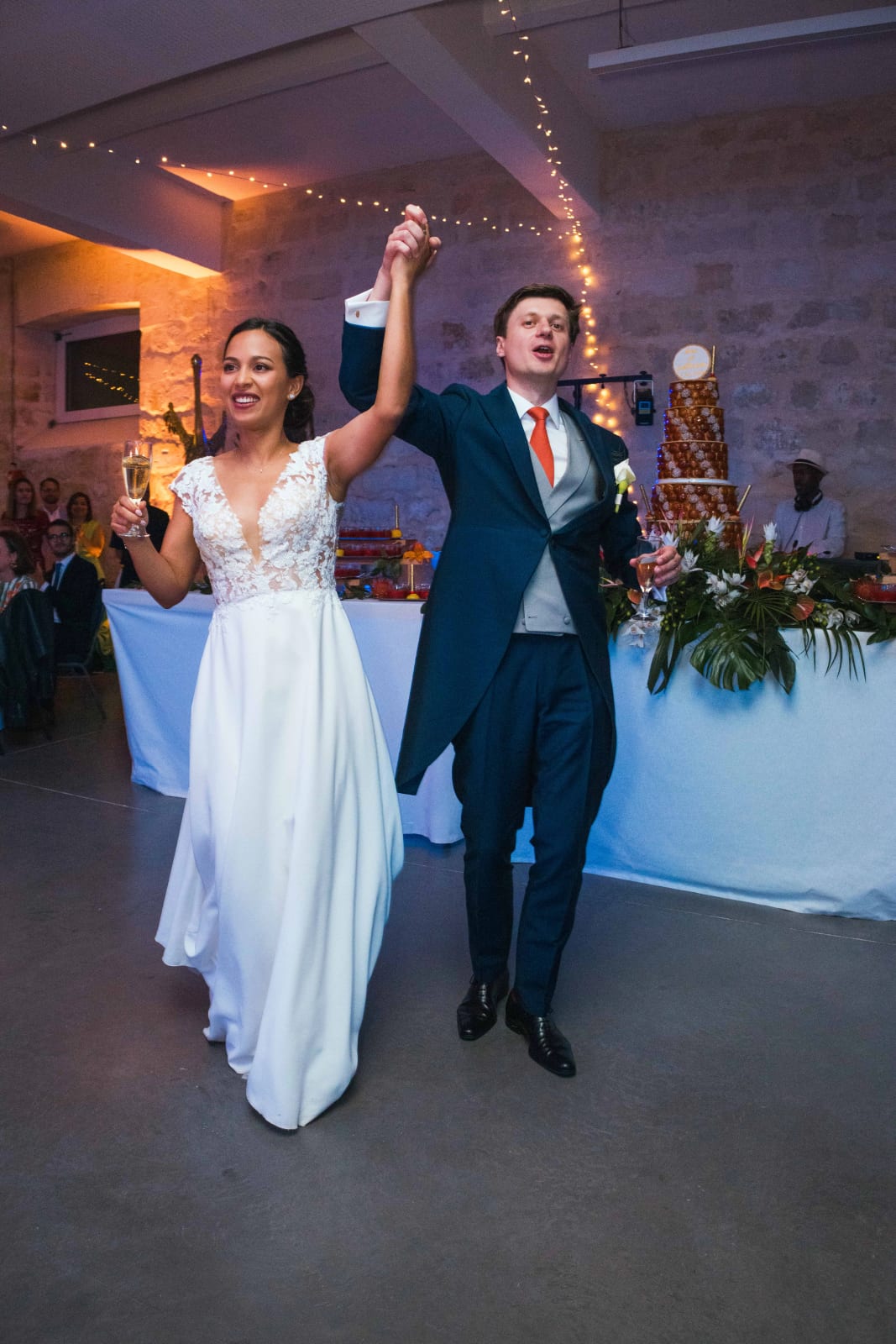 Ana et Guillaume, mains levées et coupe à la main, célèbrent devant la pièce montée sous les guirlandes lumineuses, mariage au Domaine d'Orgival, par Jean-Baptiste Argyre (Jaysphotography)