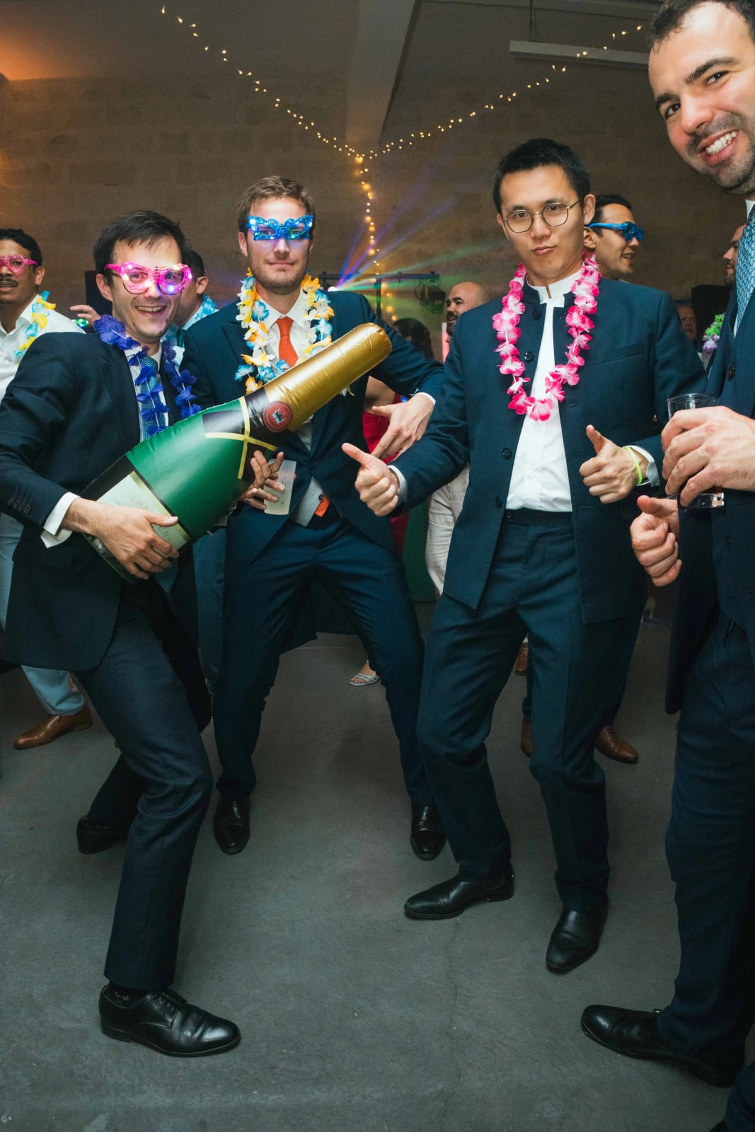 La Hora Loca : un groupe d'invités en lunettes fluo et colliers de fleurs hawaïens brandit un magnum de champagne doré, pouces levés, mariage au Domaine d'Orgival, par Jean-Baptiste Argyre (Jaysphotography)