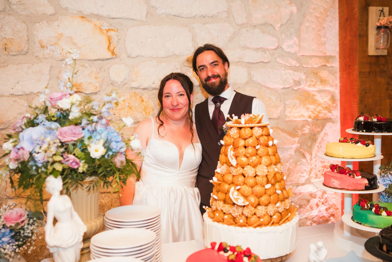 Jessie et Aurélien devant le croquembouche et les desserts au mur de pierre, mariage au Domaine de l'Orangerie de Villette, par Jean-Baptiste Argyre (Jaysphotography)