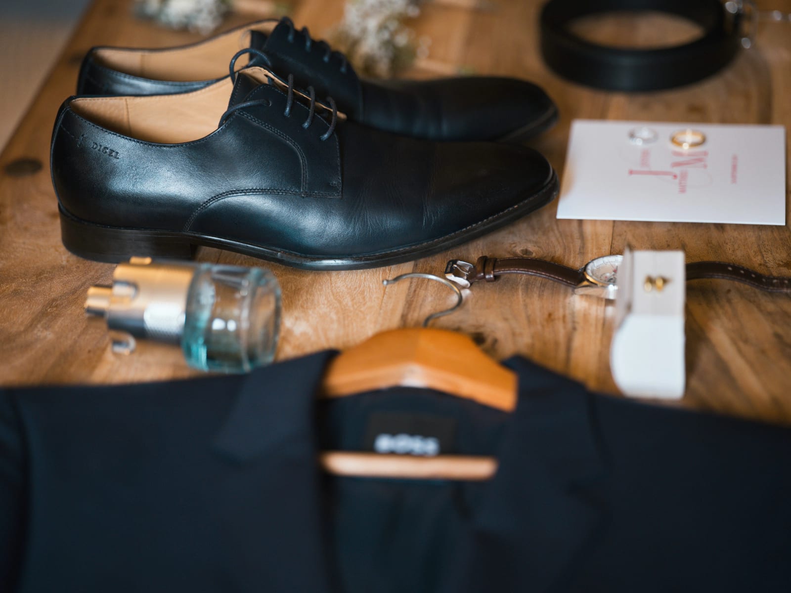 Accessoires du marié sur une table en bois : derbies noires, ceinture, parfum, montre, boutons de manchette et alliances posées sur un carton aux lettres roses, Domaine le Pommerieux, mariage au Domaine le Pommerieux, Buzancy, par Jean-Baptiste Argyre (Jaysphotography)
