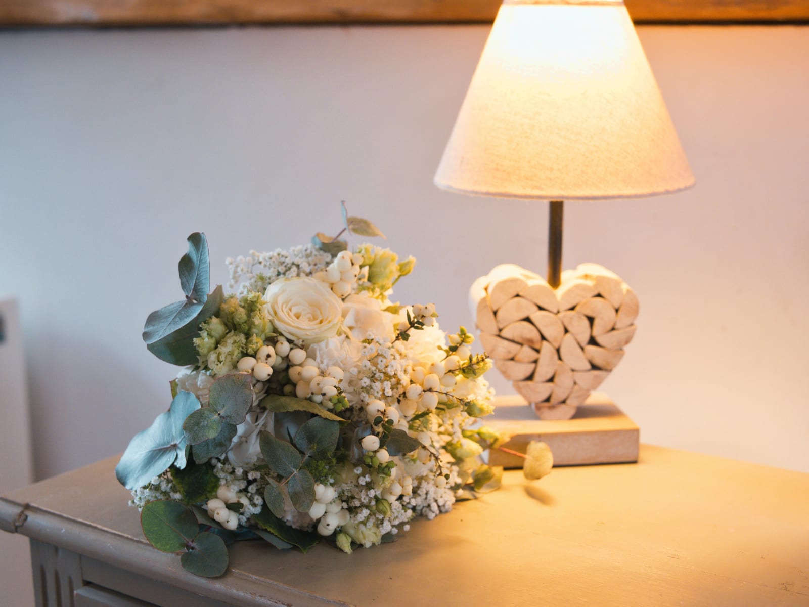 Bouquet de roses blanches, eucalyptus, gypsophile et baies blanches posé sur une commode près d'une lampe au pied en forme de cœur, mariage au Domaine le Pommerieux, Buzancy, par Jean-Baptiste Argyre (Jaysphotography)