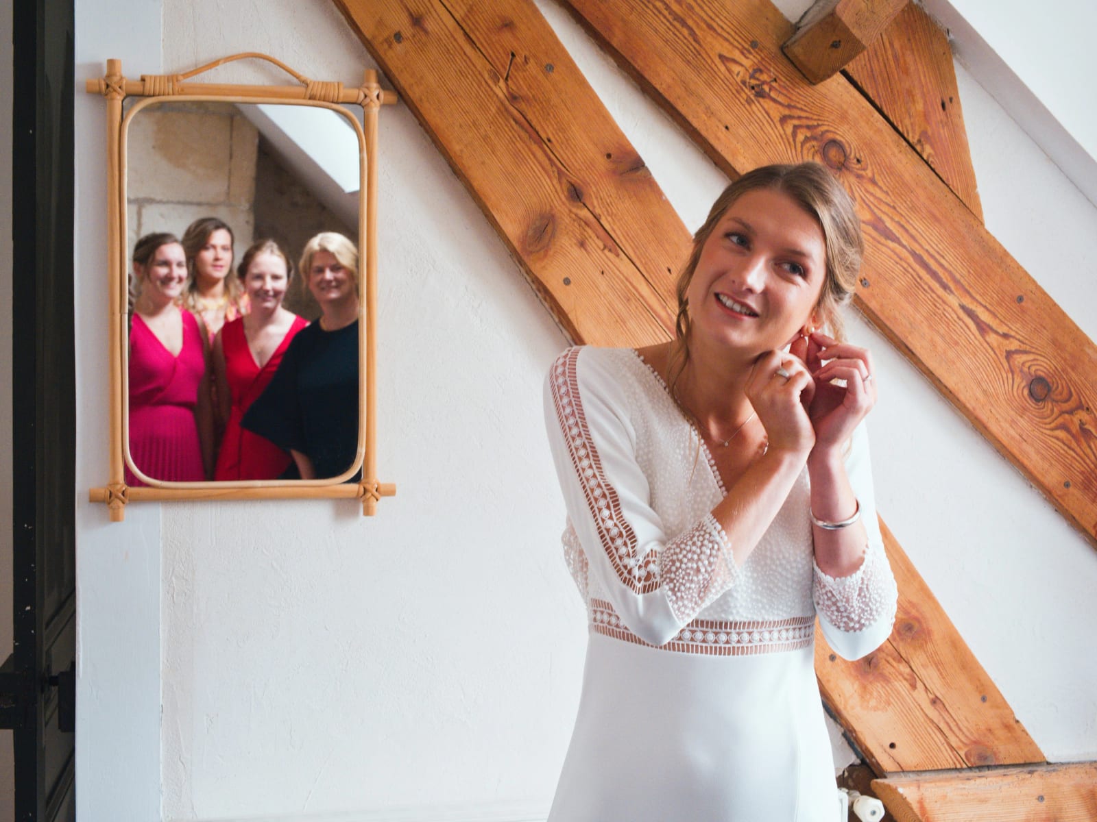 La mariée met ses boucles d'oreilles sous les poutres, quatre amies en robes roses la regardent dans un miroir de rotin, mariage au Domaine le Pommerieux, Buzancy, par Jean-Baptiste Argyre (Jaysphotography)
