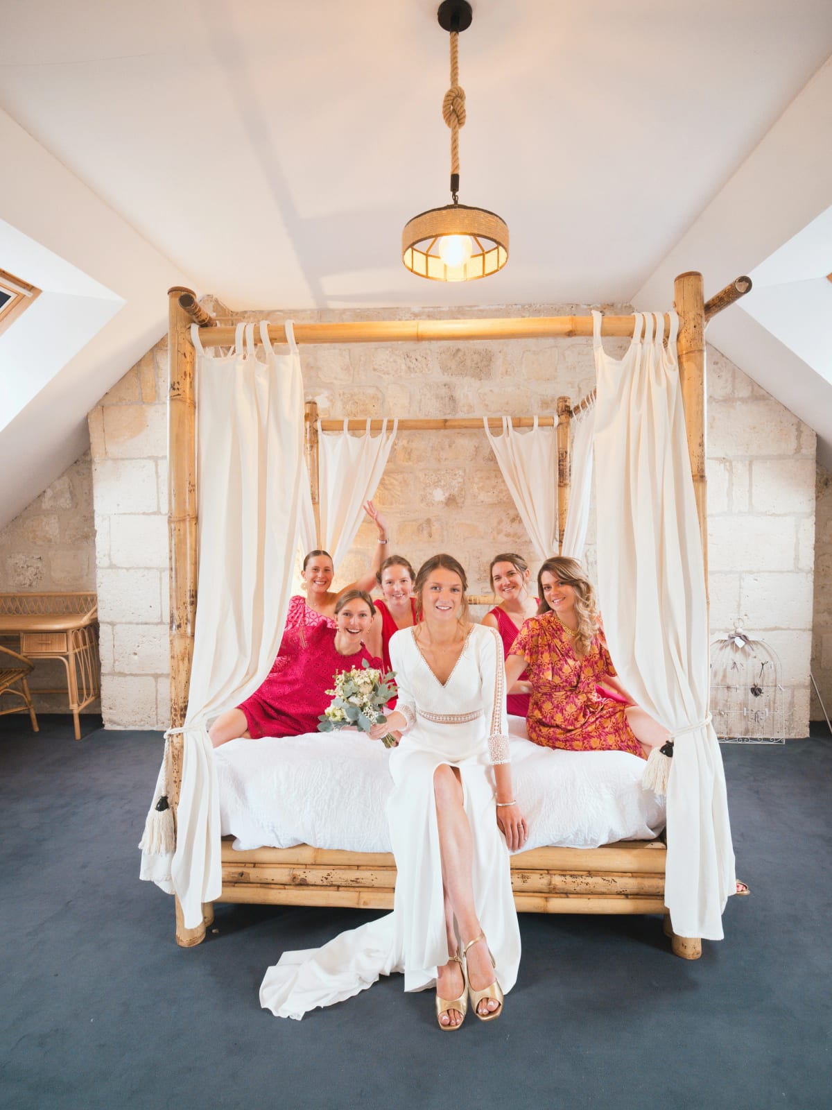 La mariée assise au pied d'un lit à baldaquin en bambou, ses amies en robes roses et framboise derrière elle, mariage au Domaine le Pommerieux, Buzancy, par Jean-Baptiste Argyre (Jaysphotography)