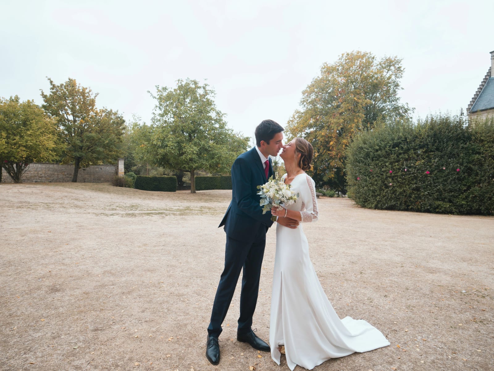 Les mariés s'embrassent au milieu d'un parc à la pelouse sèche, arbres qui commencent à jaunir, mariage au Domaine le Pommerieux, Buzancy, par Jean-Baptiste Argyre (Jaysphotography)