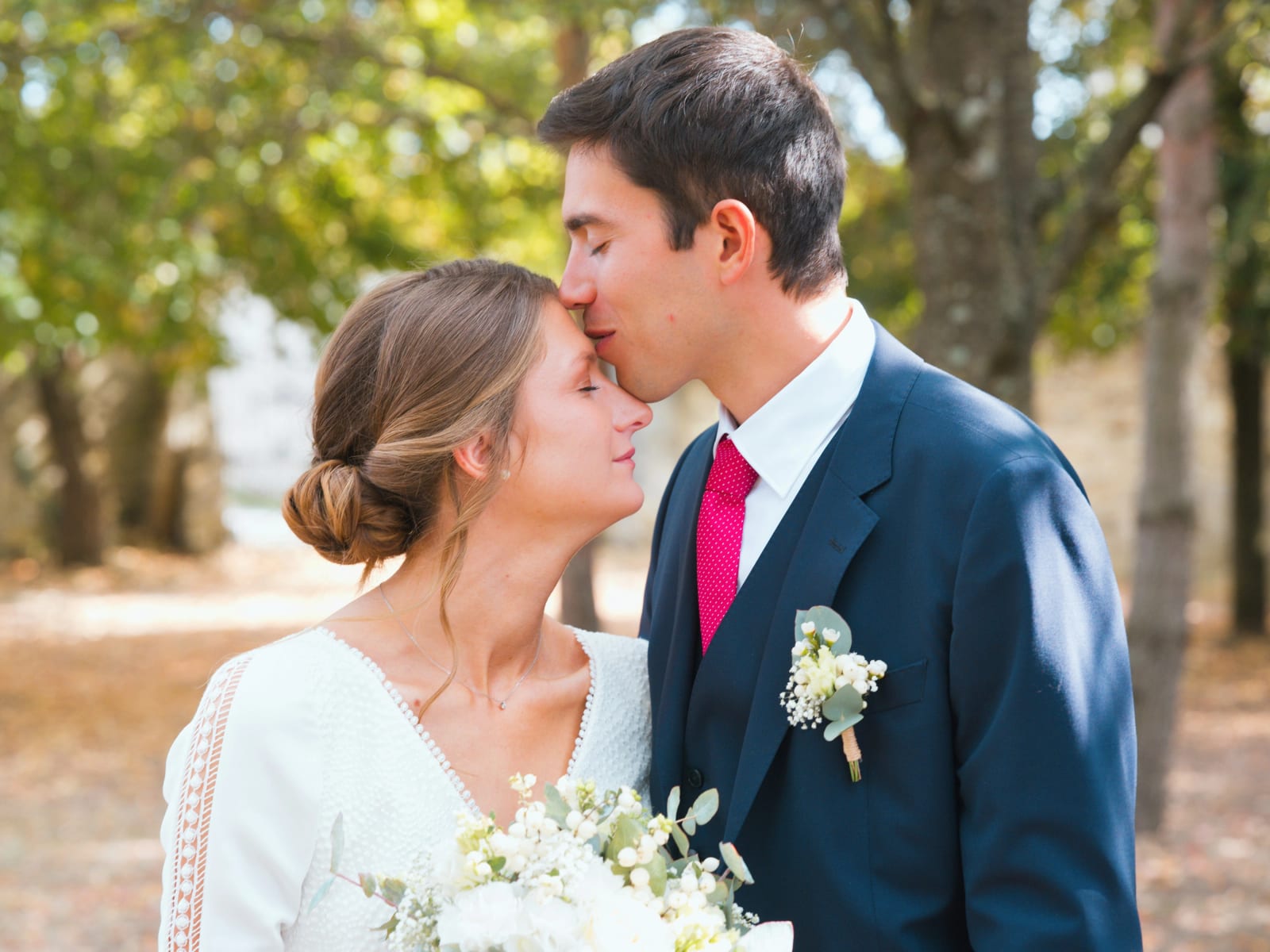 Le marié embrasse la mariée sur le front sous les arbres, bouquet blanc et boutonnière, mariage au Domaine le Pommerieux, Buzancy, par Jean-Baptiste Argyre (Jaysphotography)