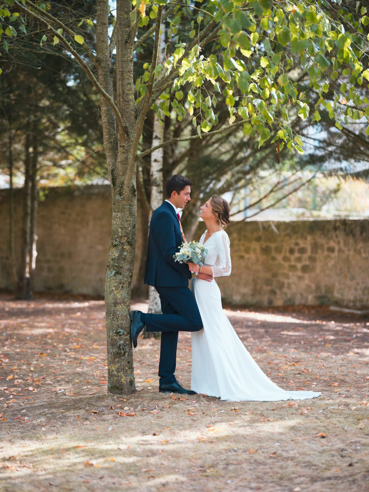 Le marié adossé à un arbre, un pied contre le tronc, enlace la mariée devant un mur de pierre, mariage au Domaine le Pommerieux, Buzancy, par Jean-Baptiste Argyre (Jaysphotography)