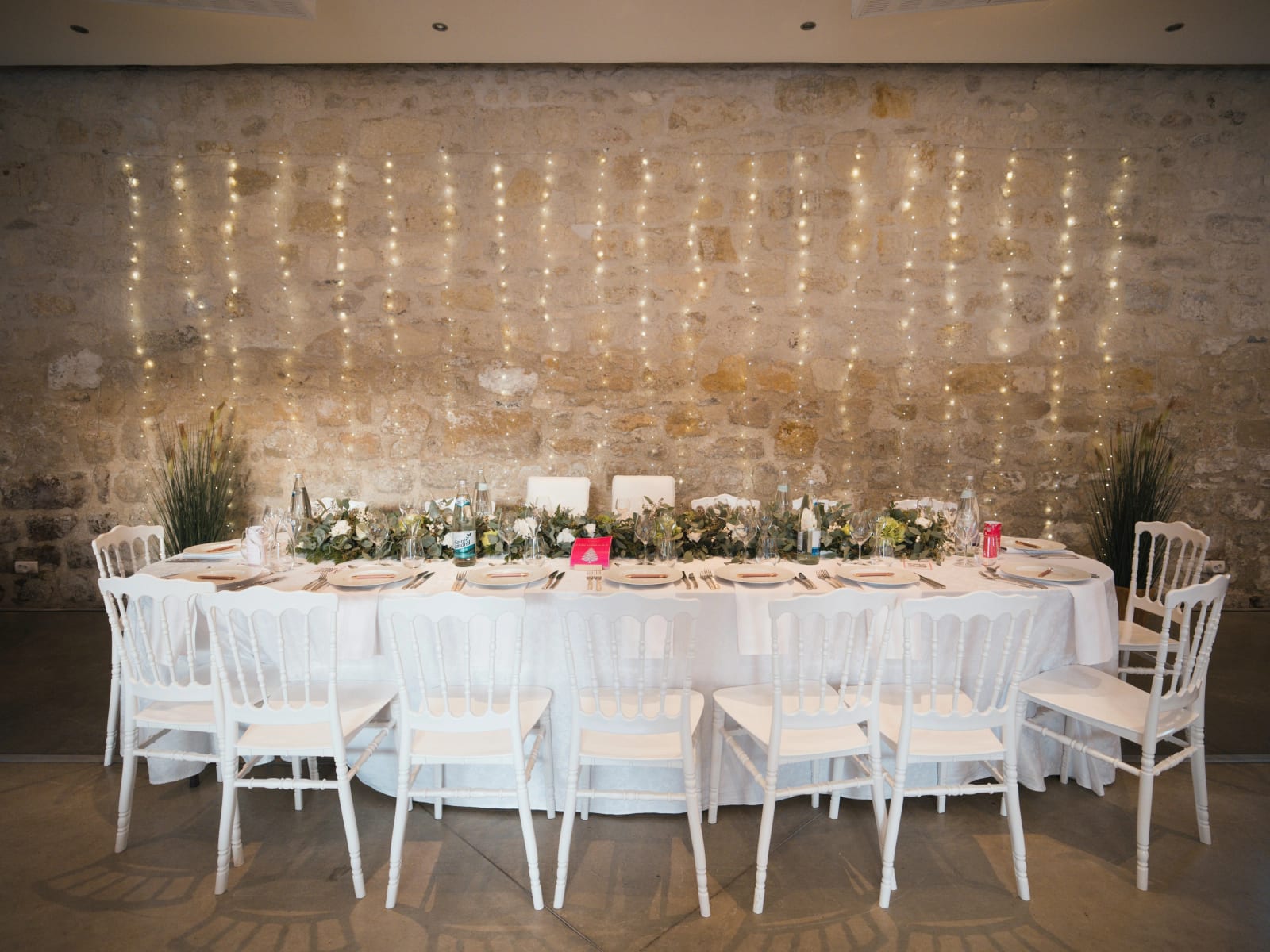 Table d'honneur nappée de blanc, chaises Napoléon blanches et chemin d'eucalyptus devant un mur de pierre tendu de guirlandes lumineuses, mariage au Domaine le Pommerieux, Buzancy, par Jean-Baptiste Argyre (Jaysphotography)