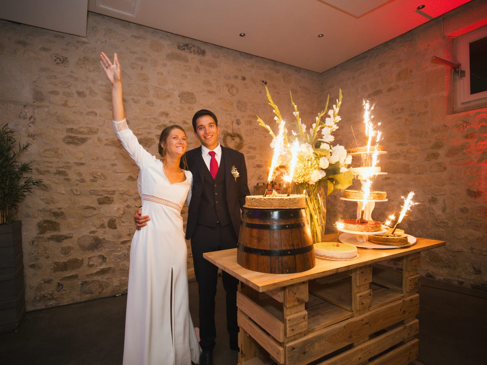 Les mariés devant le dessert aux fontaines d'étincelles : gâteau sur un tonneau et présentoir de tartes sur une table en palettes, mariage au Domaine le Pommerieux, Buzancy, par Jean-Baptiste Argyre (Jaysphotography)