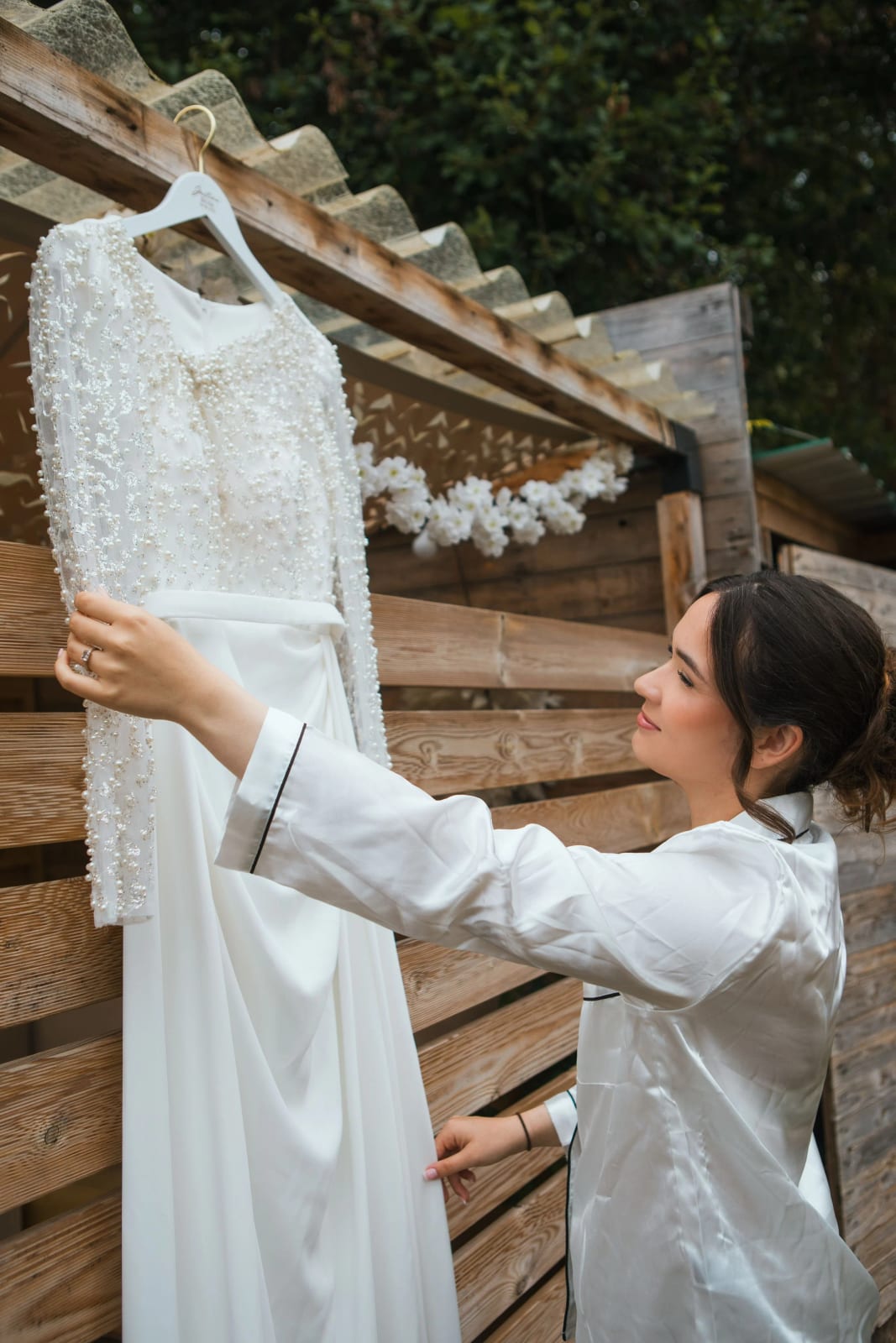 La mariée en pyjama de satin blanc accroche sa robe à manches longues brodée de perles à une palissade de bois, mariage à la Salle Georges Brassens, Villers-Saint-Paul, par Jean-Baptiste Argyre (Jaysphotography)