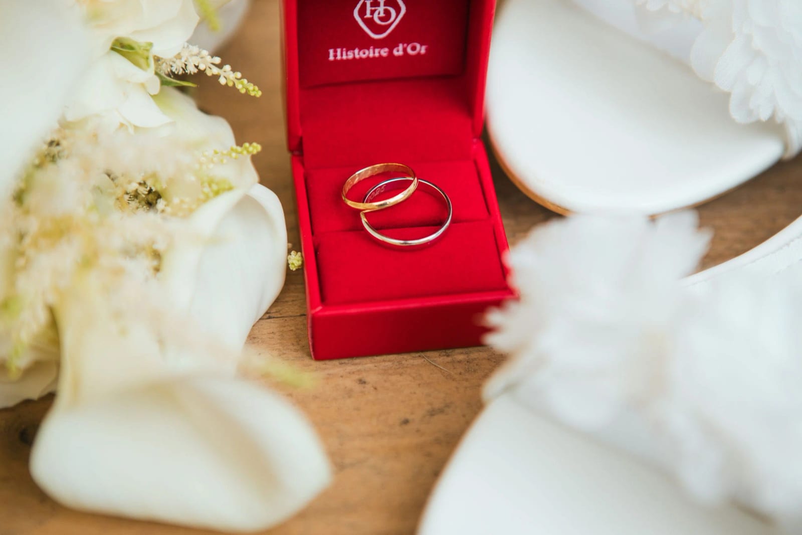 Alliances dans un écrin rouge Histoire d'Or, entre des arums blancs et des chaussons de mariée, mariage à la Salle Georges Brassens, Villers-Saint-Paul, par Jean-Baptiste Argyre (Jaysphotography)