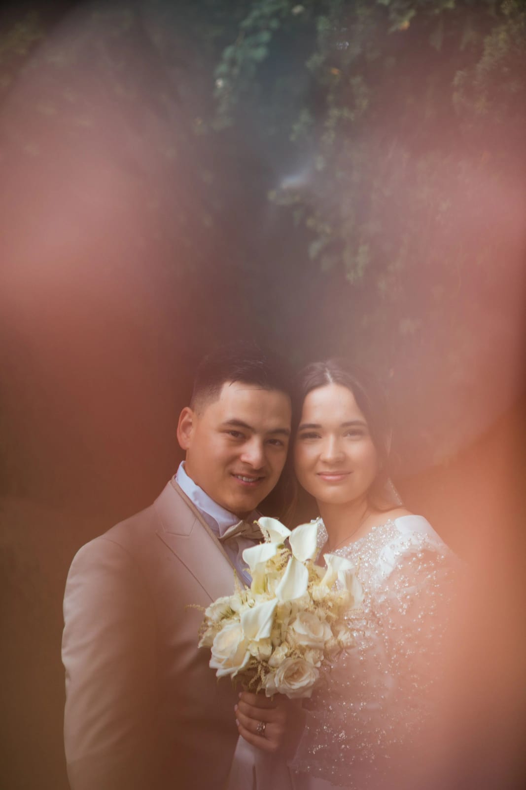 Portrait des mariés pris à travers un premier plan rose flou, bouquet d'arums blancs, mariage à la Salle Georges Brassens, Villers-Saint-Paul, par Jean-Baptiste Argyre (Jaysphotography)