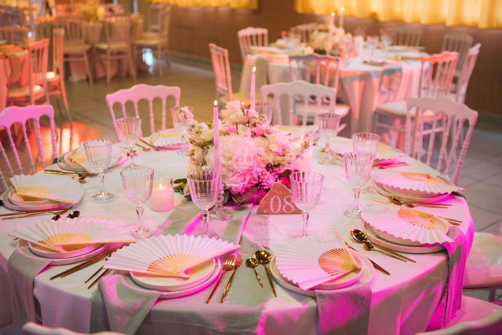 Table ronde dressée sous lumière rose à la Salle Georges Brassens : éventails plissés, couverts dorés, bougies et centre fleuri, mariage à la Salle Georges Brassens, Villers-Saint-Paul, par Jean-Baptiste Argyre (Jaysphotography)
