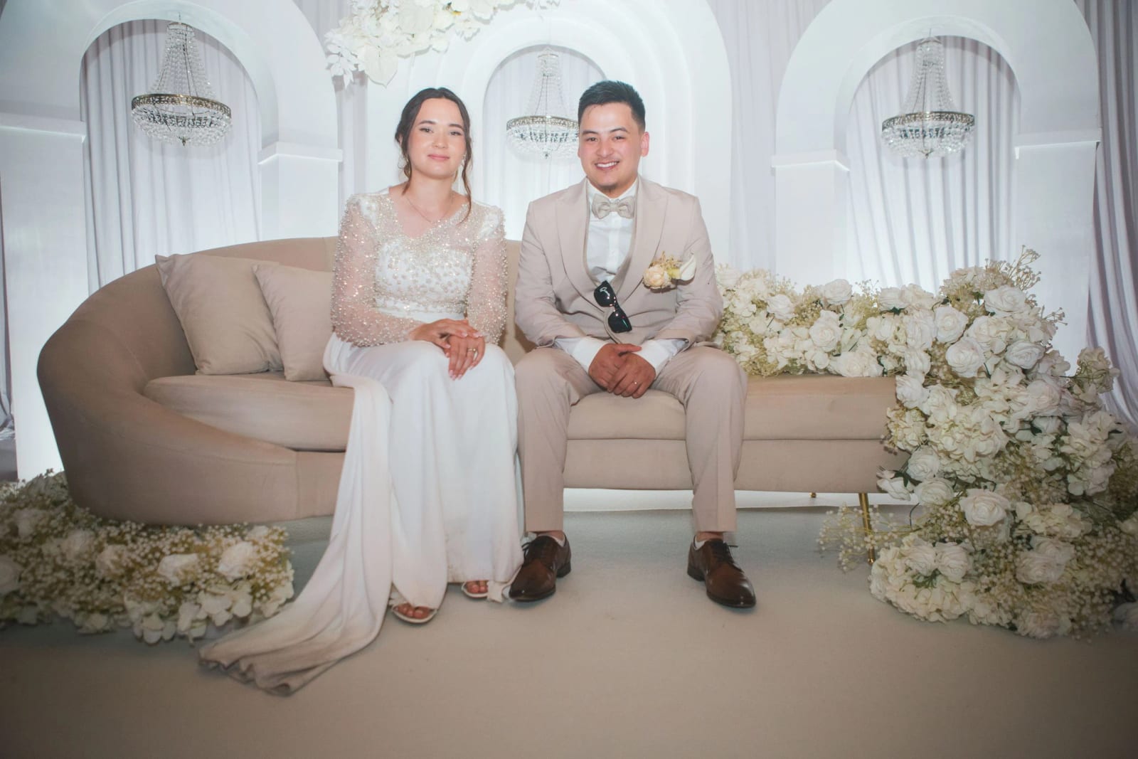 Les mariés assis sur un canapé arrondi sous des arches blanches et des lustres, massifs de fleurs blanches, mariage à la Salle Georges Brassens, Villers-Saint-Paul, par Jean-Baptiste Argyre (Jaysphotography)