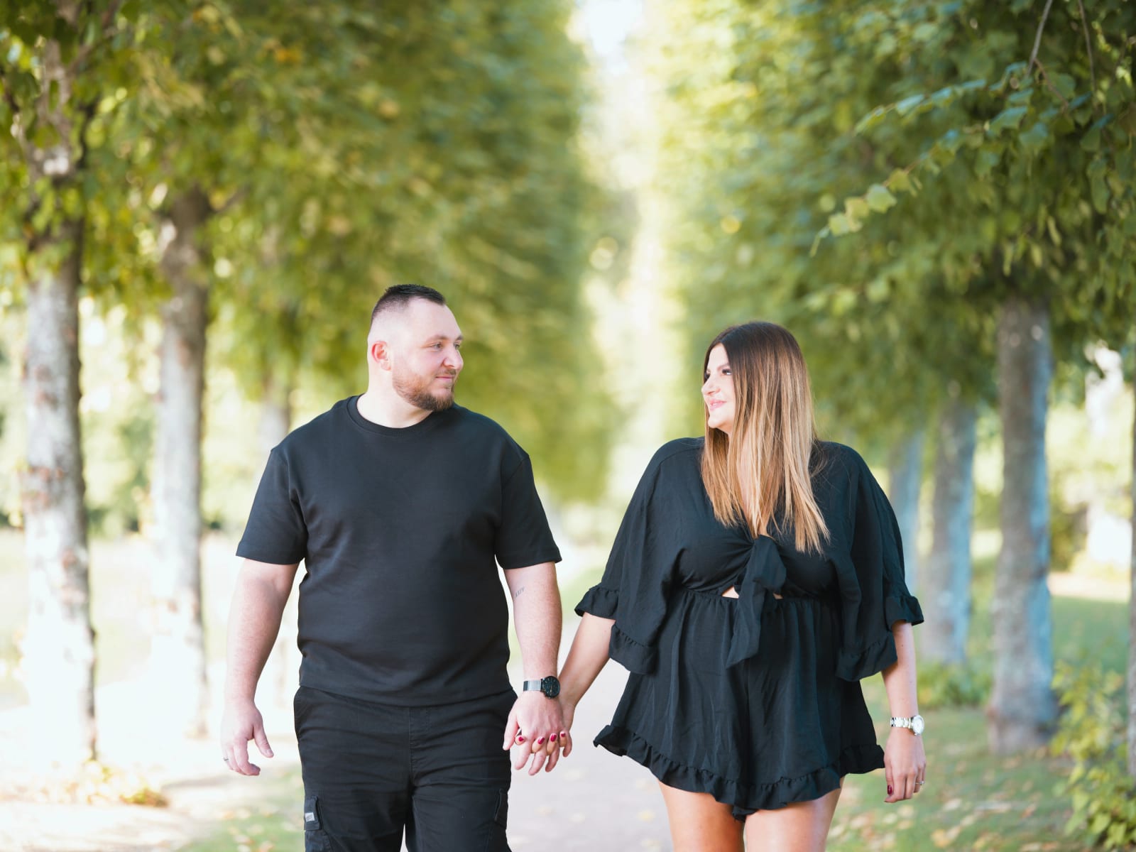 Laurena et Anthony marchent main dans la main dans une allée de tilleuls du Domaine de Villarceaux, tous deux habillés en noir, mariage au Domaine de Villarceaux, Chaussy (Val-d'Oise), par Jean-Baptiste Argyre (Jaysphotography)