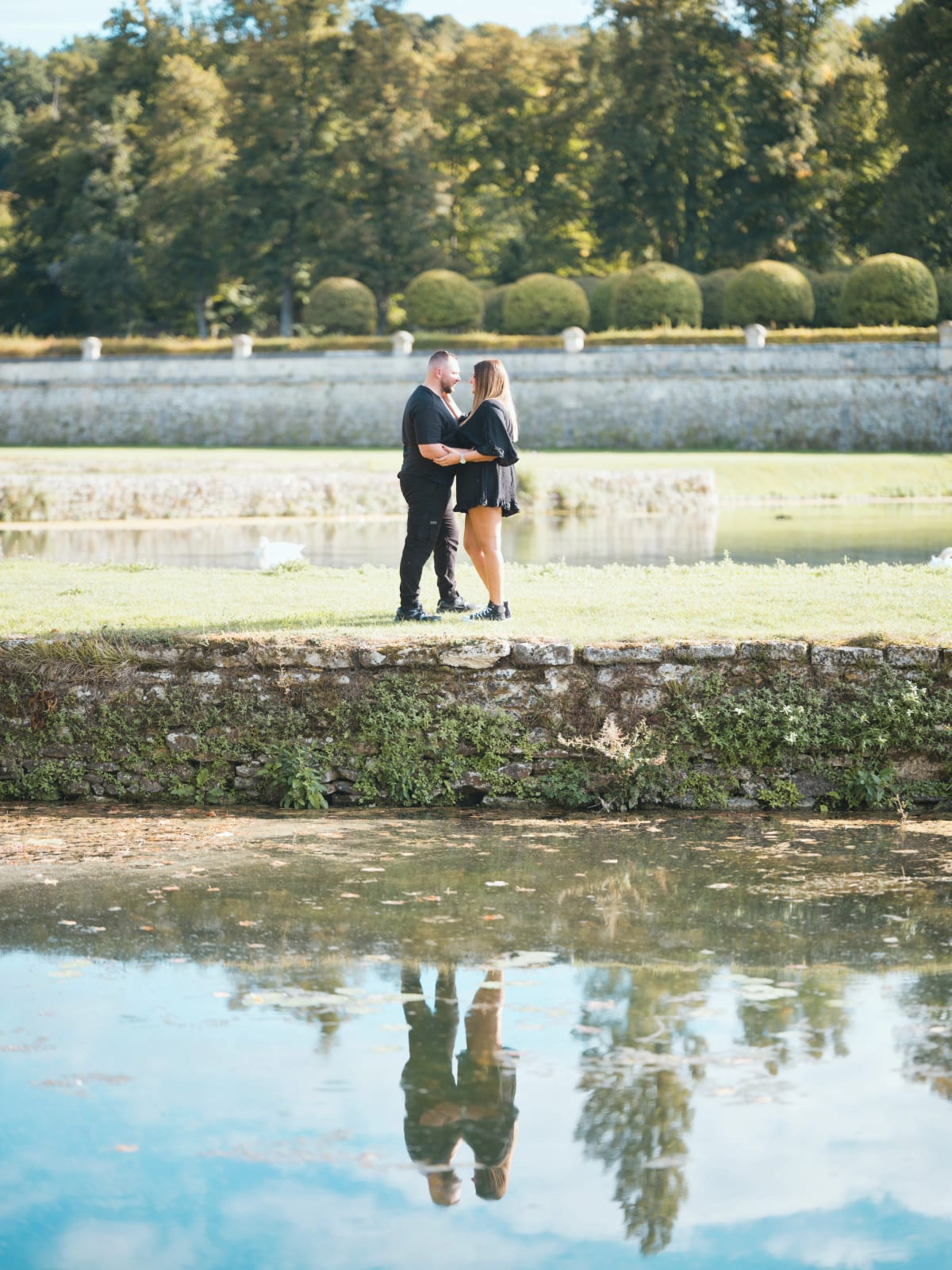 Séance pré-wedding au Domaine de Villarceaux : le couple enlacé entre deux canaux, mur de pierre et buis taillés en boule, reflet dans l'eau, par Jean-Baptiste Argyre (Jaysphotography)