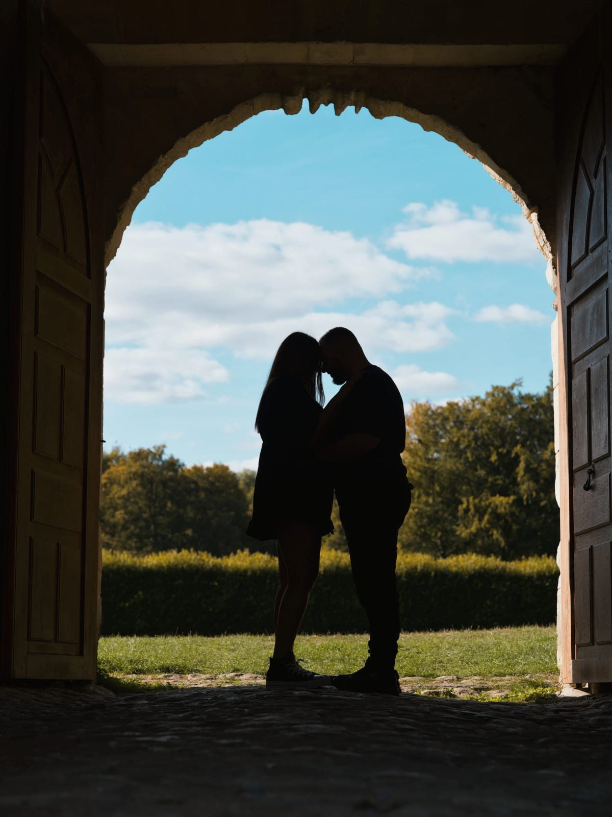 Silhouette de Laurena et Anthony front contre front sous un porche voûté aux vantaux de bois ouverts sur le ciel, mariage au Domaine de Villarceaux, Chaussy (Val-d'Oise), par Jean-Baptiste Argyre (Jaysphotography)