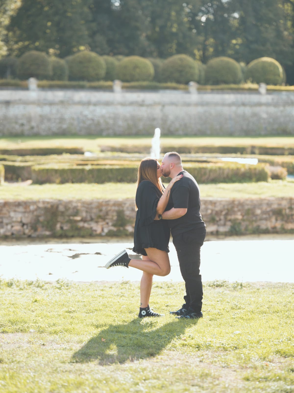 Baiser de Laurena et Anthony devant un jardin de buis et un jet d'eau, elle une jambe levée, ombres allongées sur l'herbe, mariage au Domaine de Villarceaux, Chaussy (Val-d'Oise), par Jean-Baptiste Argyre (Jaysphotography)