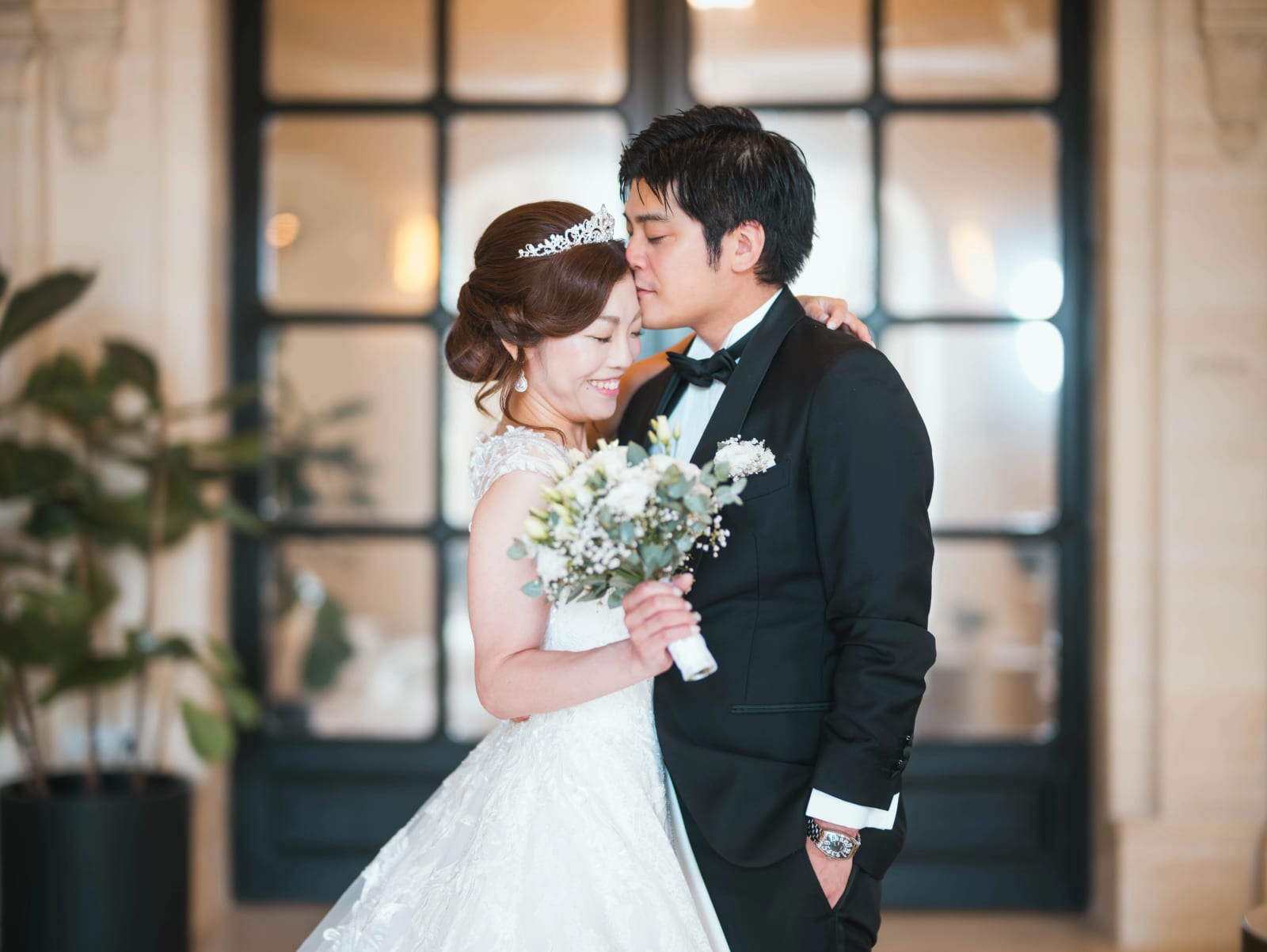 Le marié en smoking embrasse la mariée sur le front devant une porte vitrée à montants noirs, bouquet de fleurs blanches et eucalyptus, mariage au Château Mont Royal, par Jean-Baptiste Argyre (Jaysphotography)