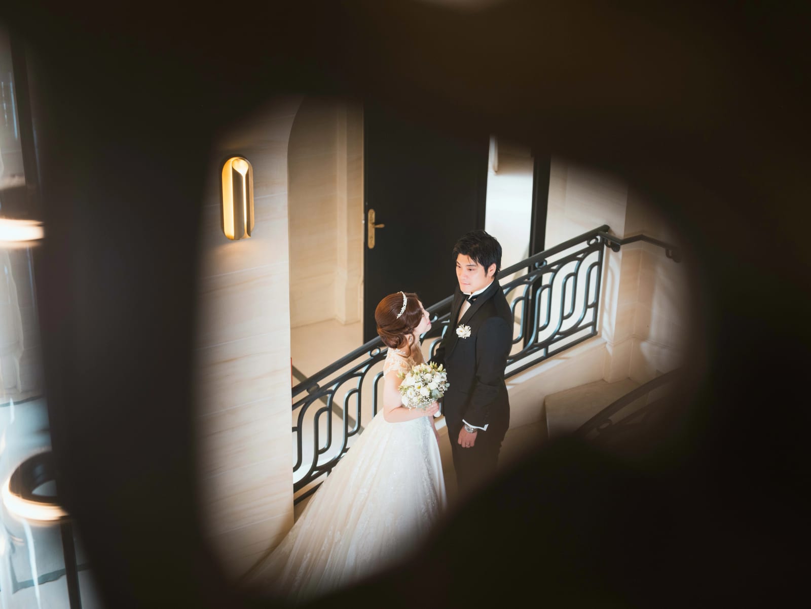 Les mariés face à face sur l'escalier à rampe de fer forgé, photographiés à travers une ouverture sombre au premier plan, mariage au Château Mont Royal, par Jean-Baptiste Argyre (Jaysphotography)