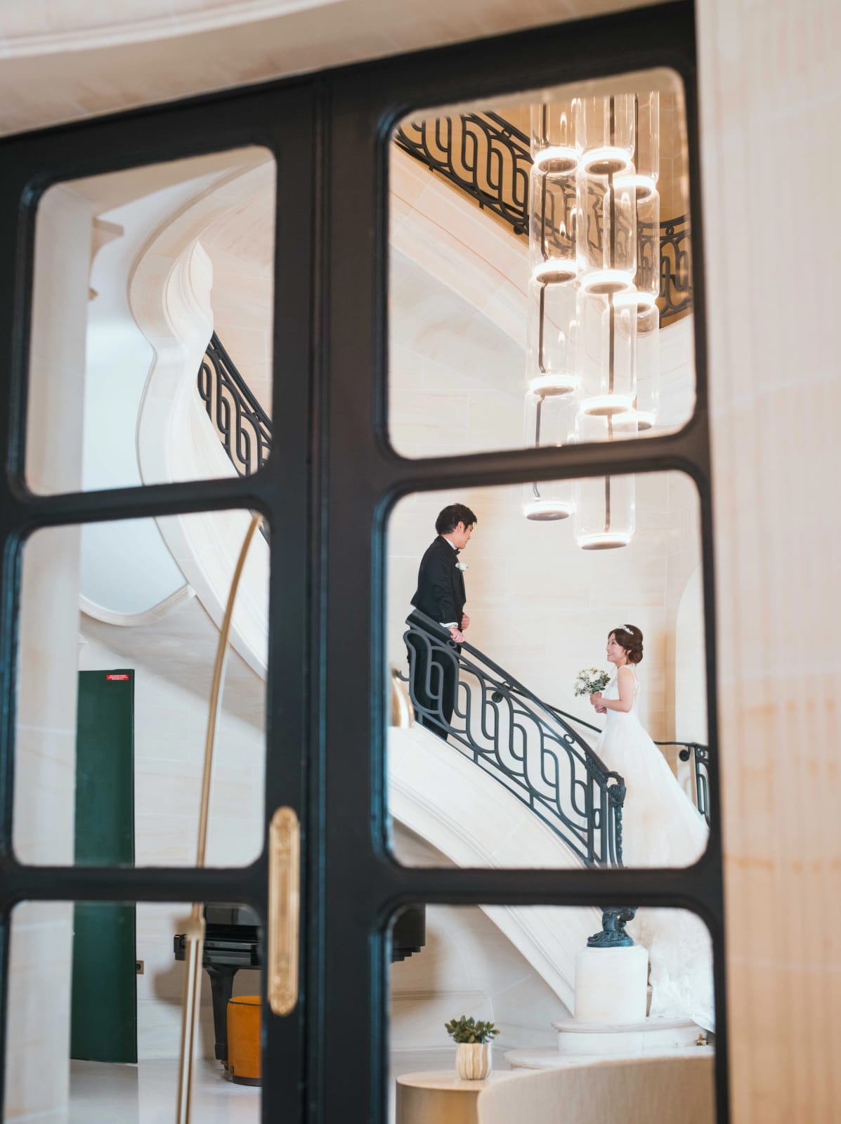 À travers les carreaux d'une porte vitrée, le marié sur les marches et la mariée qui monte l'escalier vers lui, mariage au Château Mont Royal, par Jean-Baptiste Argyre (Jaysphotography)