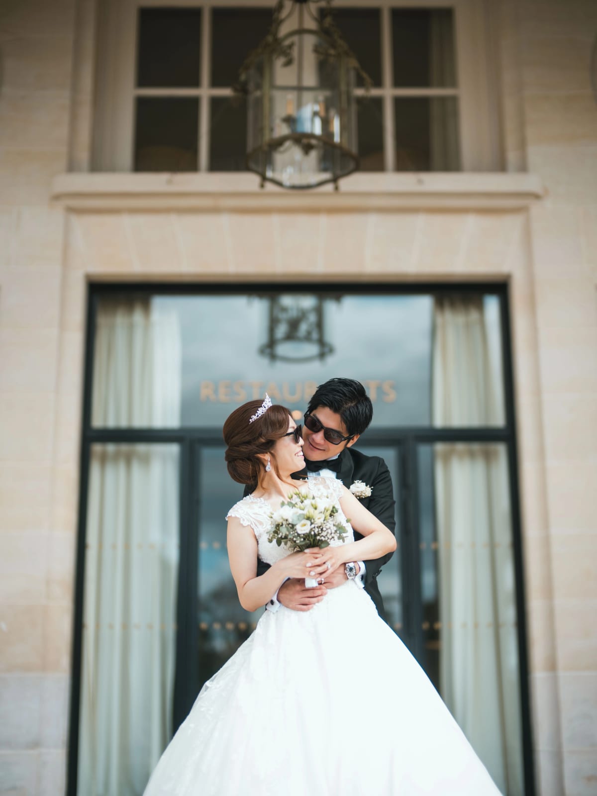 Le marié en lunettes de soleil serre la mariée par derrière devant la porte vitrée des restaurants, mariage au Château Mont Royal, par Jean-Baptiste Argyre (Jaysphotography)