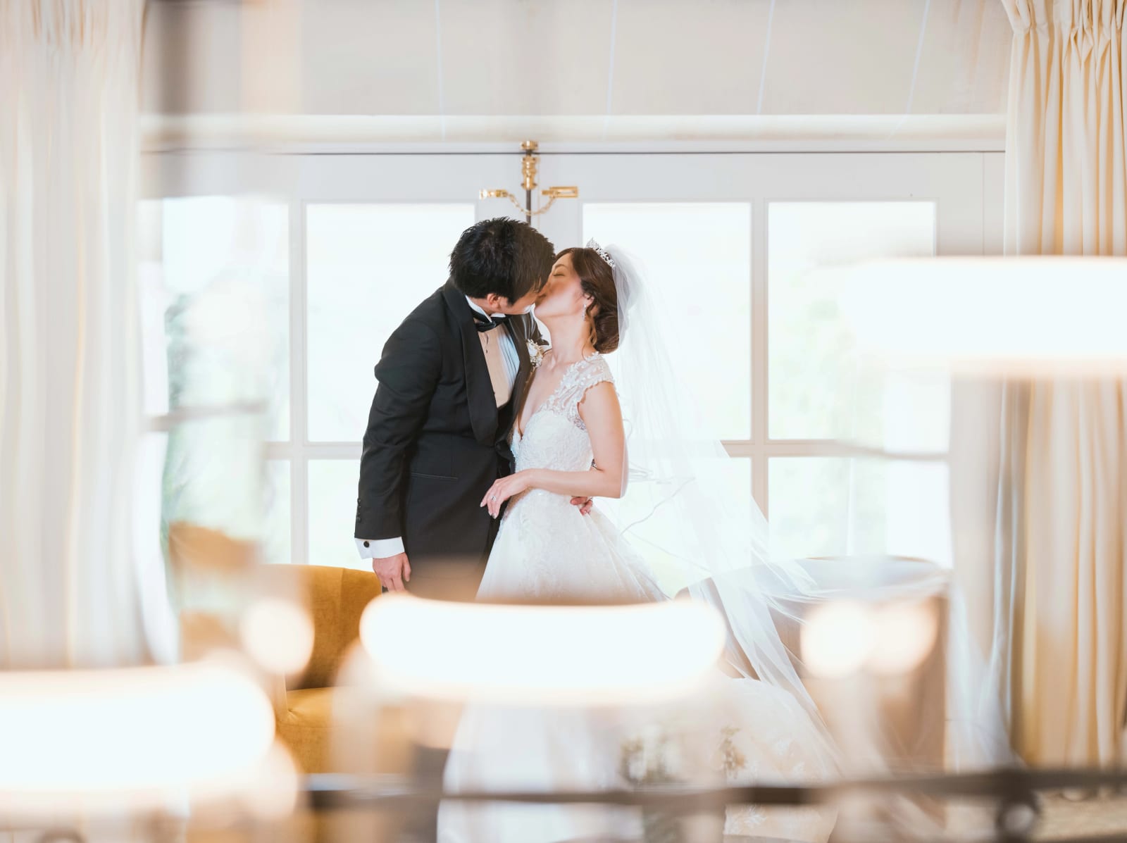 Les mariés s'embrassent devant de grandes fenêtres, la mariée en voile, lumières floues au premier plan, mariage au Château Mont Royal, par Jean-Baptiste Argyre (Jaysphotography)