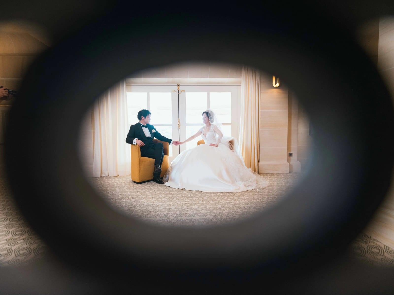 Le marié assis dans un fauteuil jaune tient la main de la mariée assise près des fenêtres, vu à travers un anneau sombre, mariage au Château Mont Royal, par Jean-Baptiste Argyre (Jaysphotography)