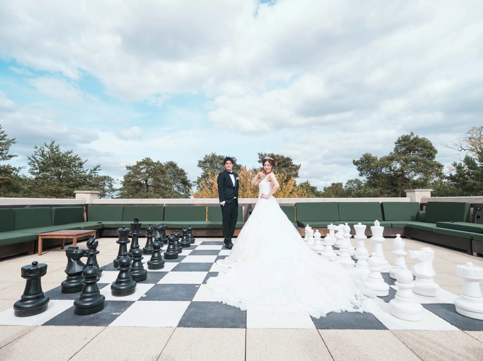 Les mariés debout sur un échiquier géant sur un toit-terrasse, banquettes vertes, pins et ciel nuageux, mariage au Château Mont Royal, par Jean-Baptiste Argyre (Jaysphotography)